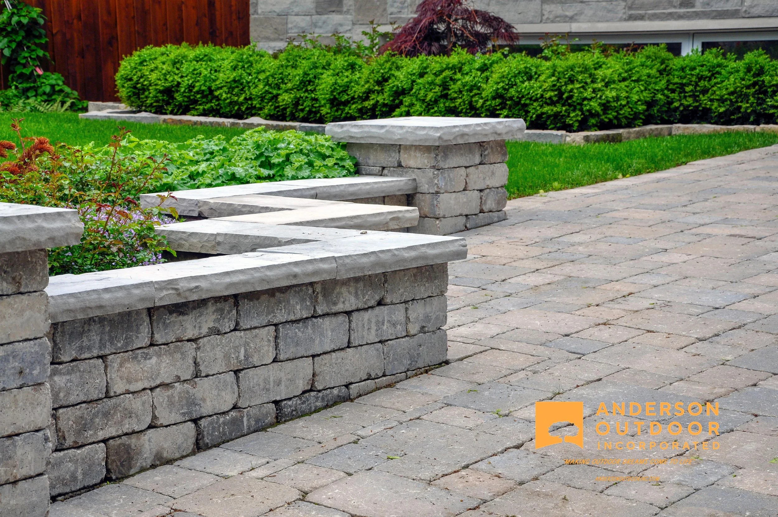 A landscaped backyard with stone retaining wall, stone and brick benches, green bushes, a small tree, and a grassy lawn. The bottom right corner has an orange logo with text reading 'Anderson Outdoor Incorporated' and the tagline 'Making outdoor drea
