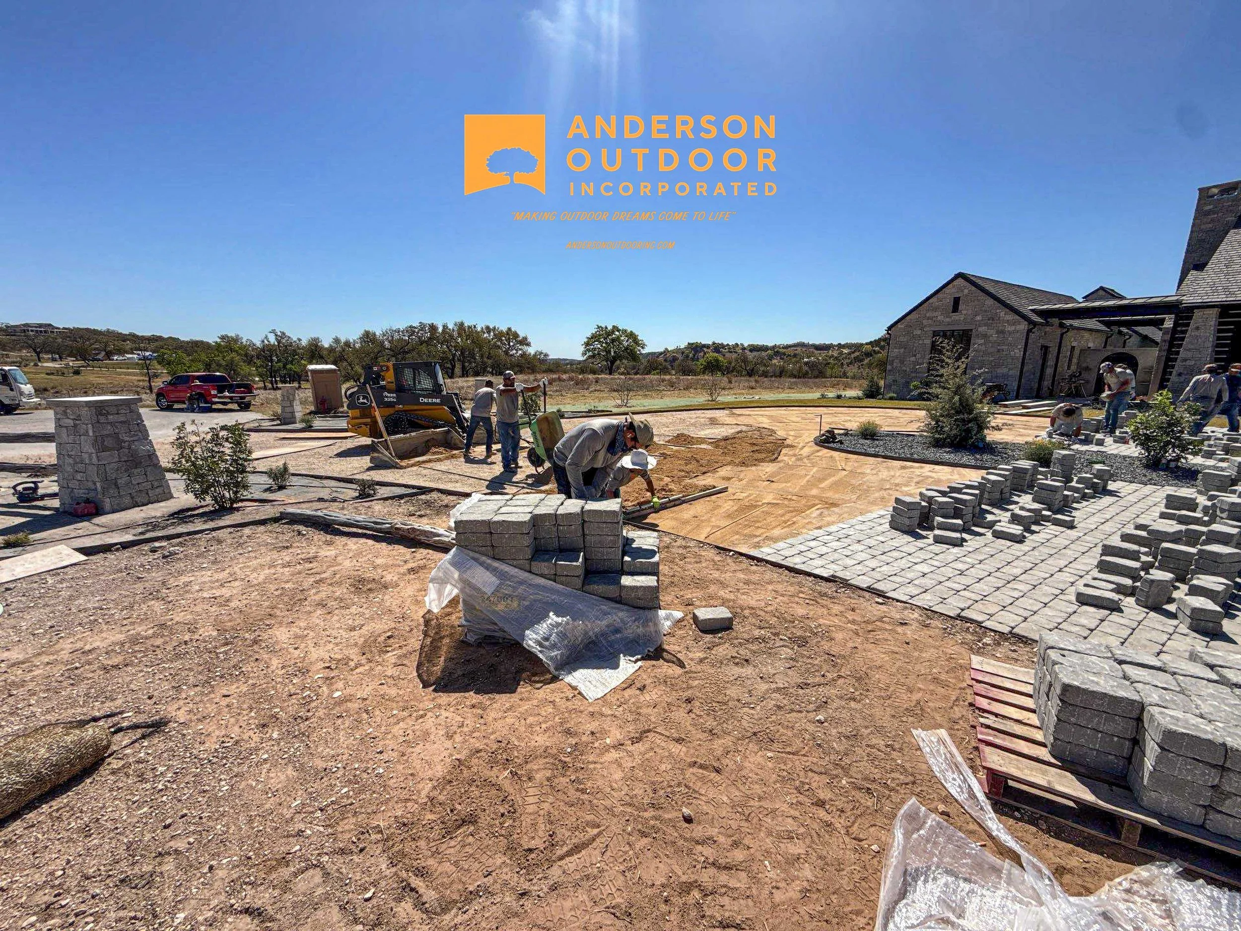 Construction workers laying paving stones on a backyard pathway, with some equipment and materials visible, under a clear blue sky.