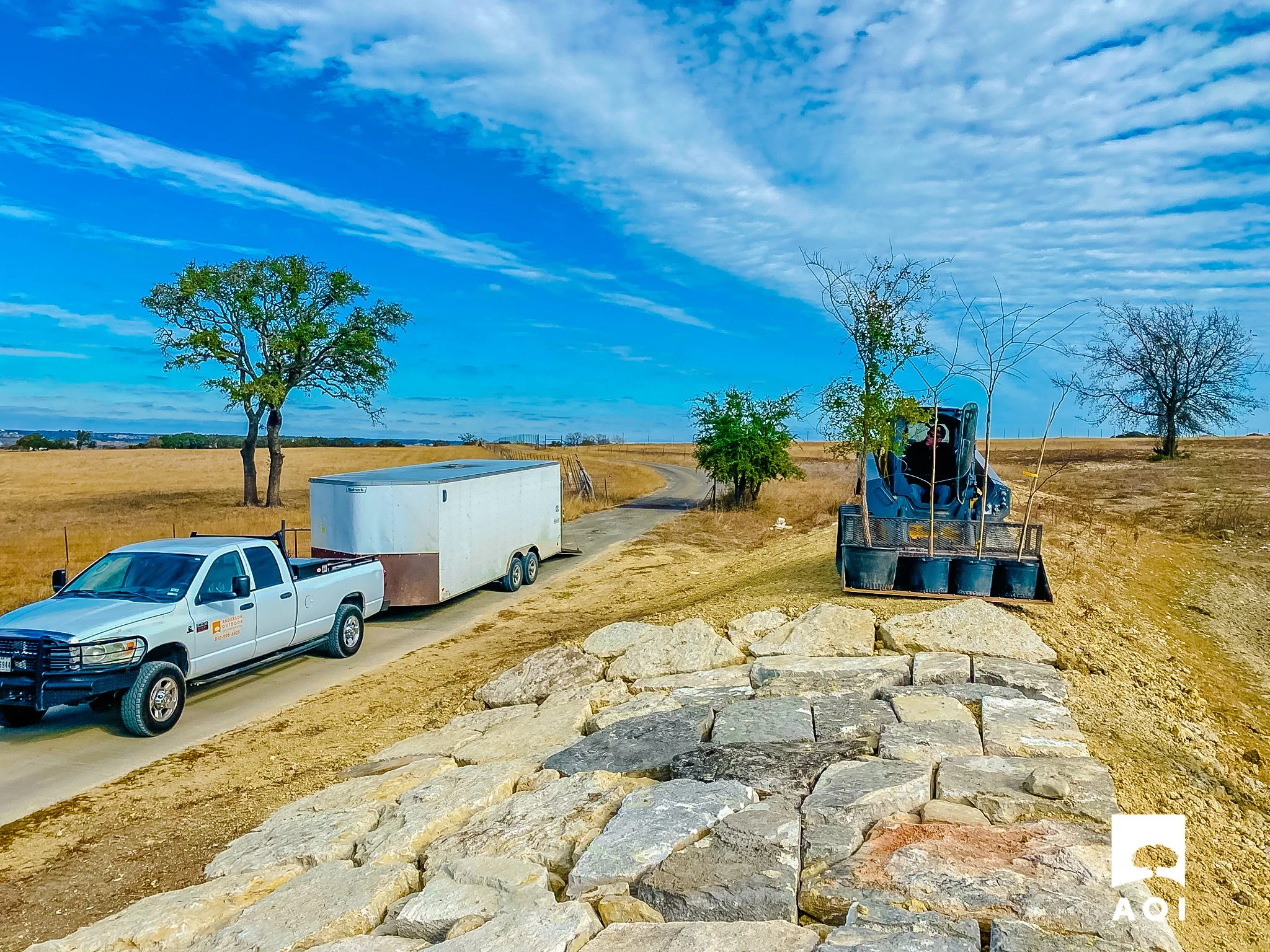 Construction vehicle and a pickup truck with a trailer on a dirt road in a rural area with trees and cloudy sky