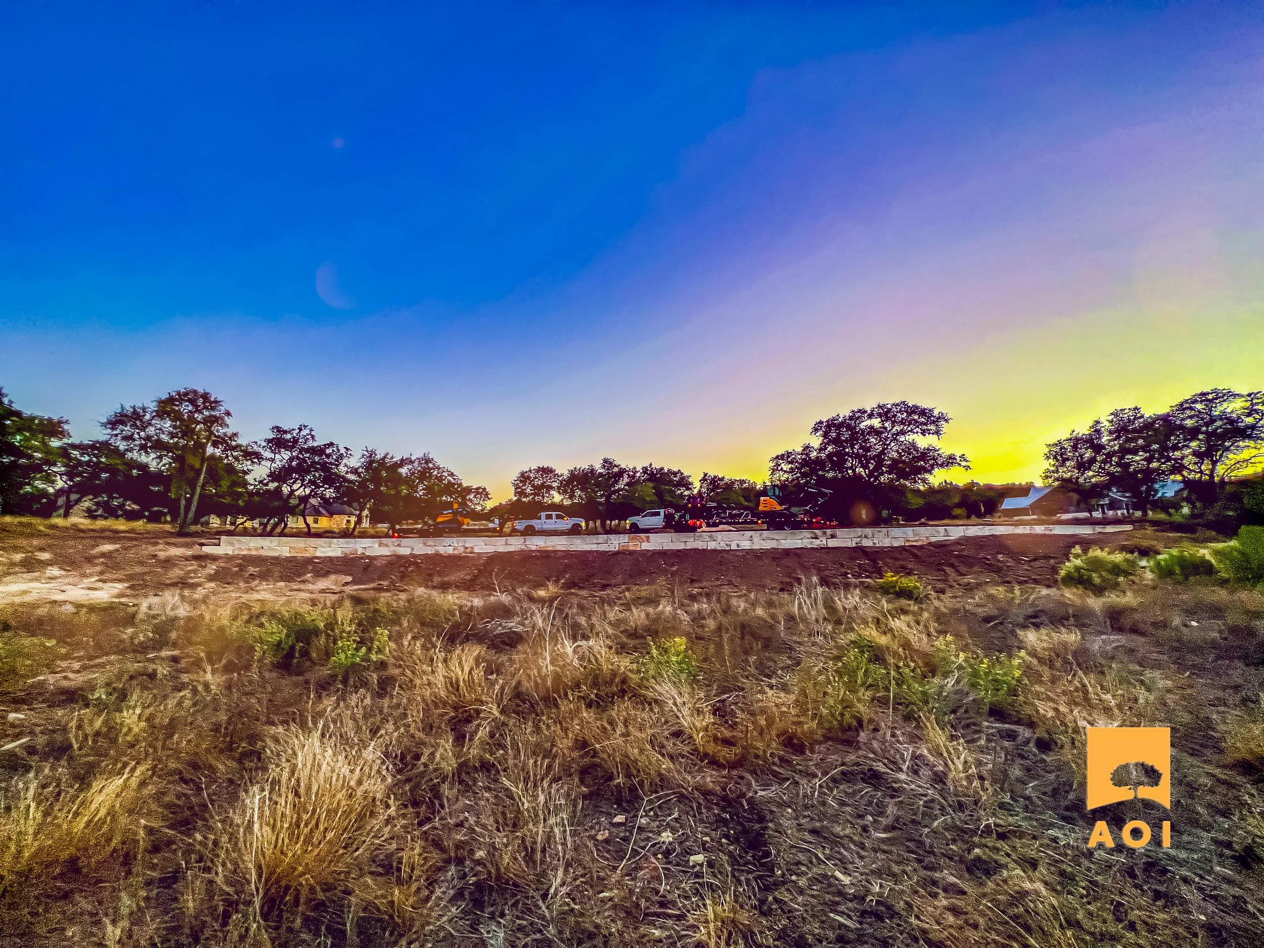 A sunset or sunrise over a rural area with trees, houses, and parked cars along the horizon, with dry grass and soil in the foreground.