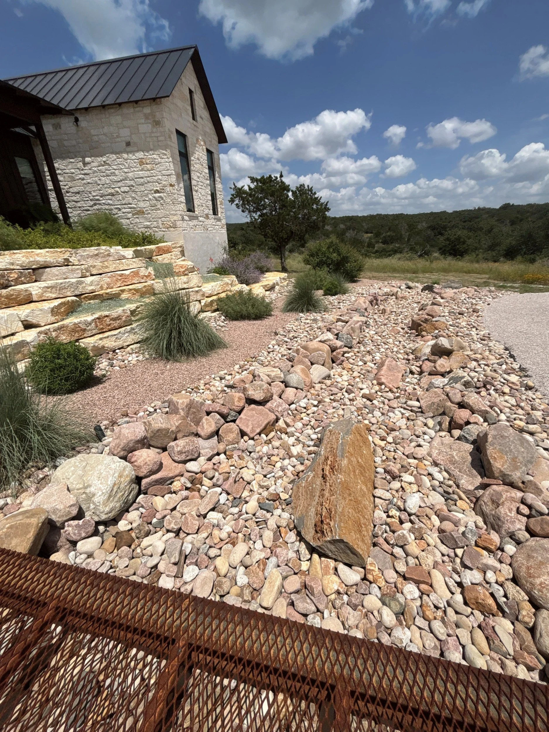 Steel grate bridge over a river rock dry creek bed beside a limestone home.