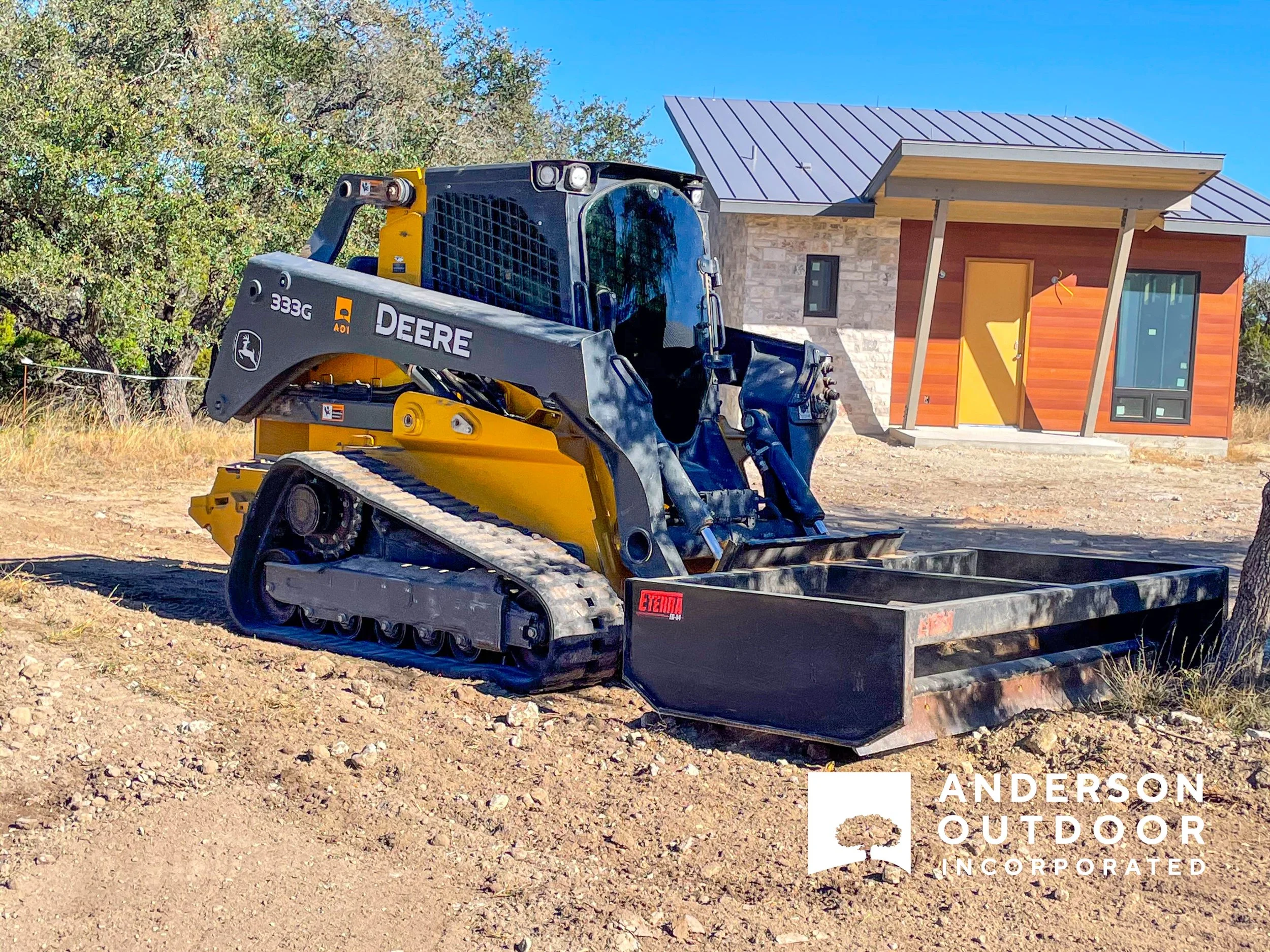 A construction site shows a small John Deere bulldozer in front of a modern house with a metal roof and wooden siding, under a clear blue sky.