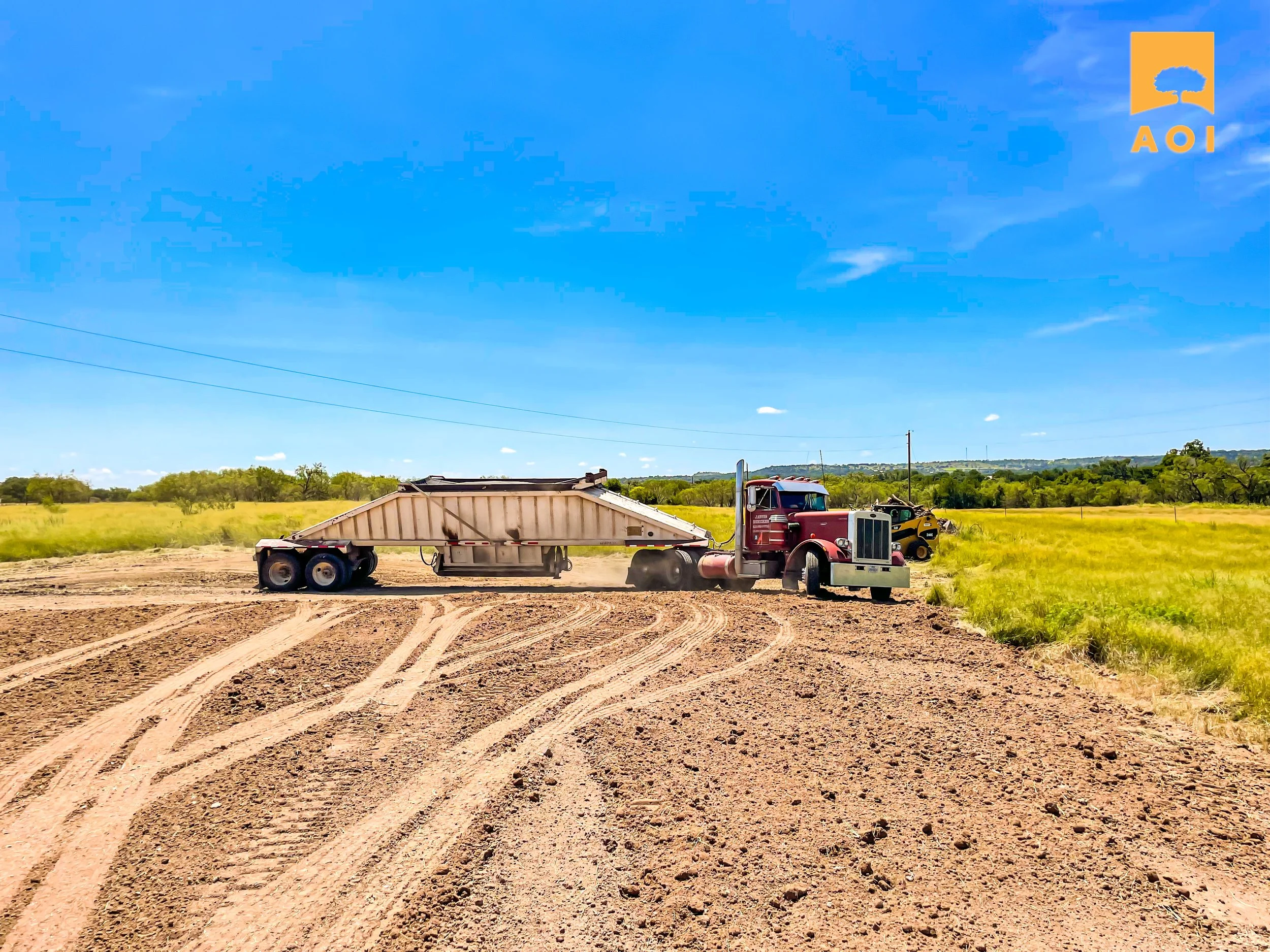 A red semi-truck pulling a flatbed trailer driving on a dirt field with tire tracks, dirt dust, and green grass in the background under a blue sky with some clouds.