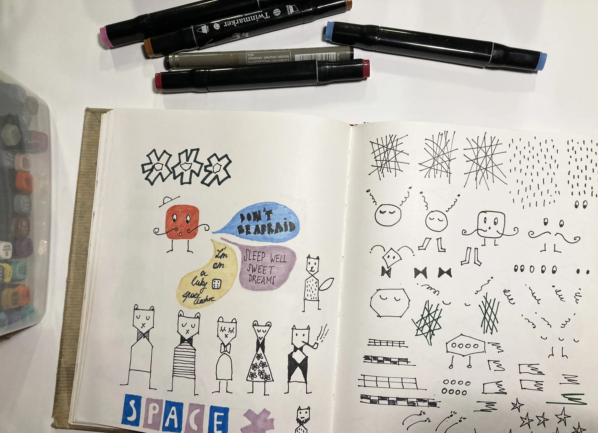 Open sketchbook with colorful and black ink doodles, including cats, flowers, abstract patterns, and motivational phrases, on a white surface with markers and pens nearby.