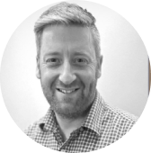 Black and white photo of a smiling man with short, styled hair and a checkered shirt.