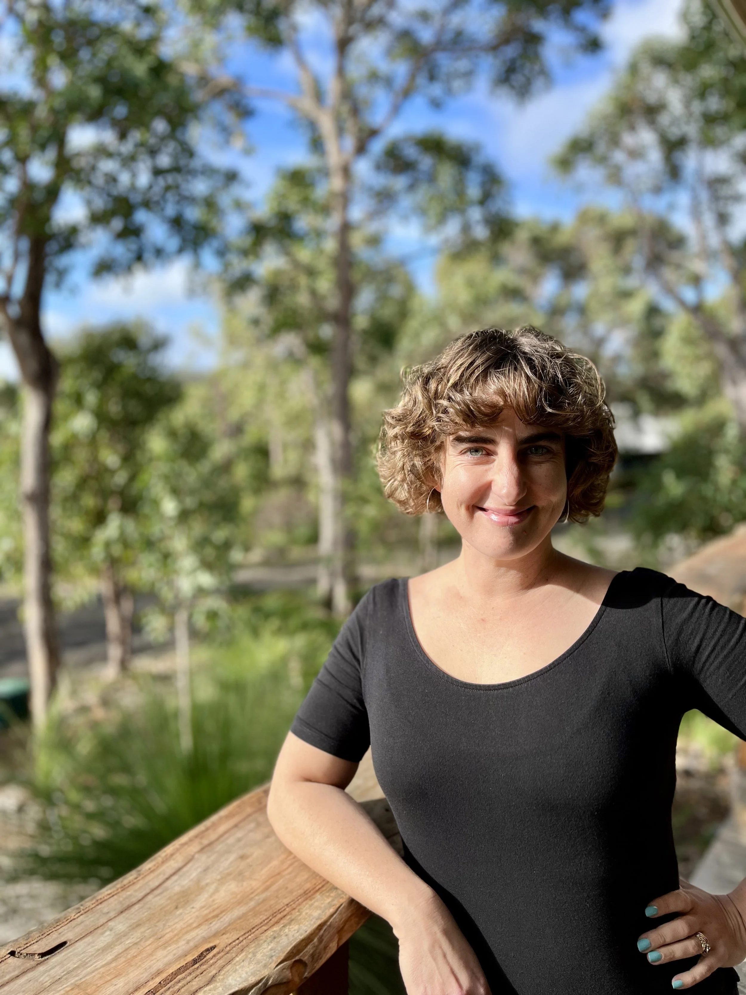 Lucienne, a naturopath in Margaret River, stands on a deck overlooking bush. Lucienne has brown, curly hair and is wearing a black top. She is smiling warmly.