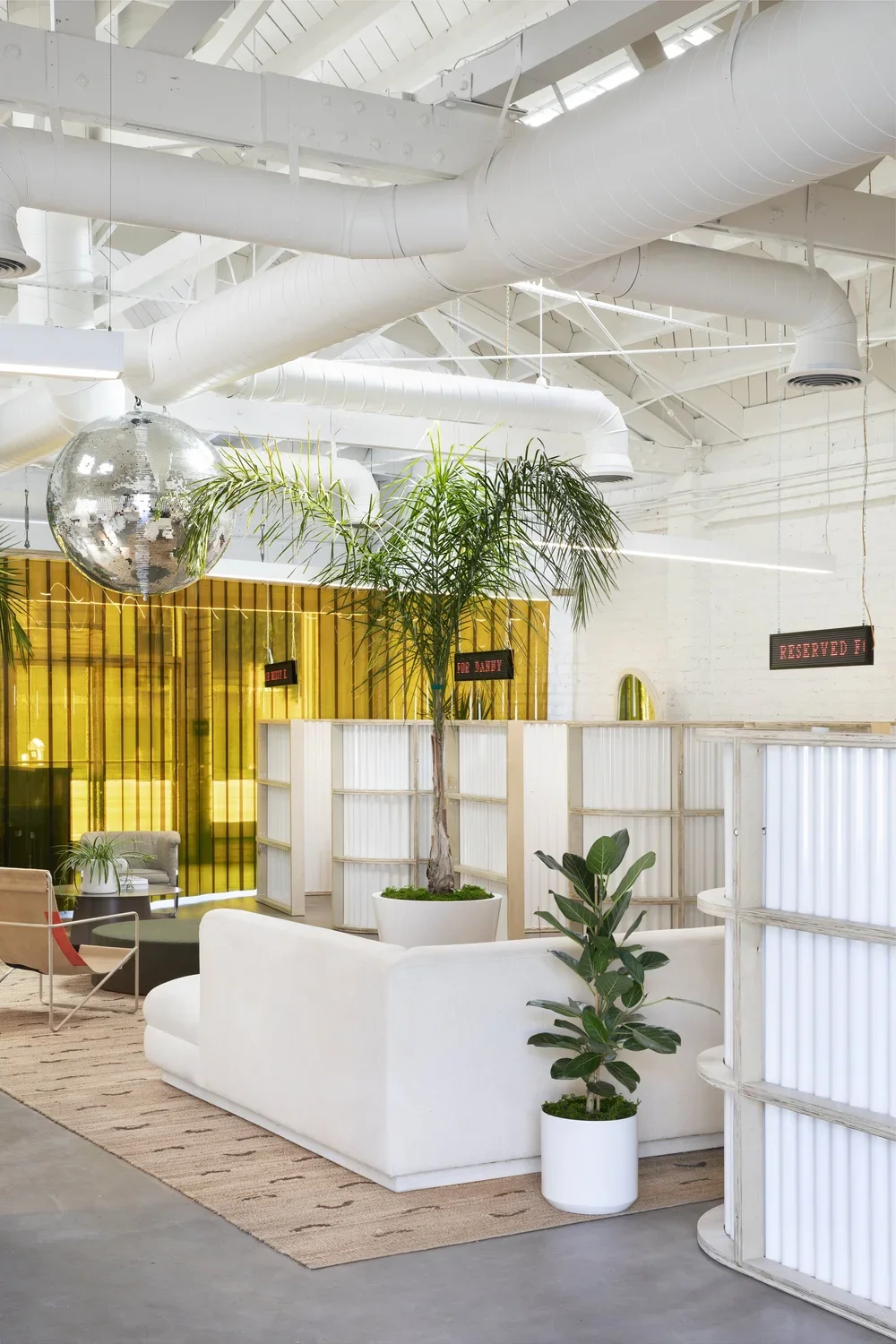 Modern indoor lounge with white furniture, potted plants, a large palm, and a disco ball, featuring overhead white pipes and a yellow beaded curtain in the background.