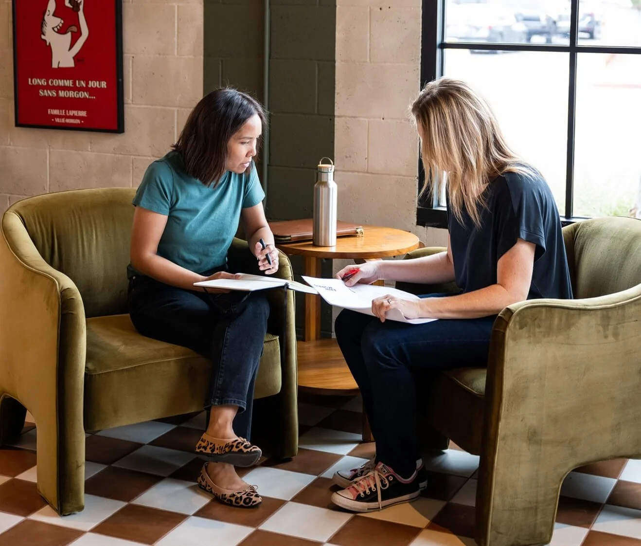 Two women sitting on green armchairs across from each other in a cafe, discussing documents. One woman has dark hair and is wearing a teal shirt and patterned shoes, holding a pen, while the other has blonde hair, is wearing a black shirt and sneakers, and is holding papers. A small wooden table with a water bottle and notebook is between them, with a window showing outside scenery.