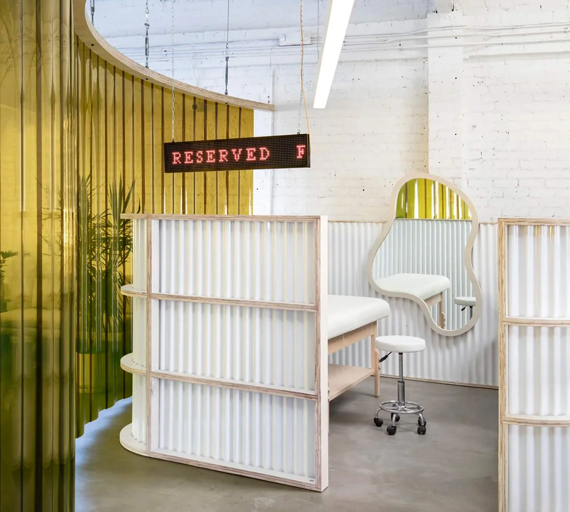 Interior of a modern space with white brick walls, yellow vertical partition, and hanging LED sign that reads "Reserved F". There is a white chair with a curved mirror in front and a rolling stool nearby.