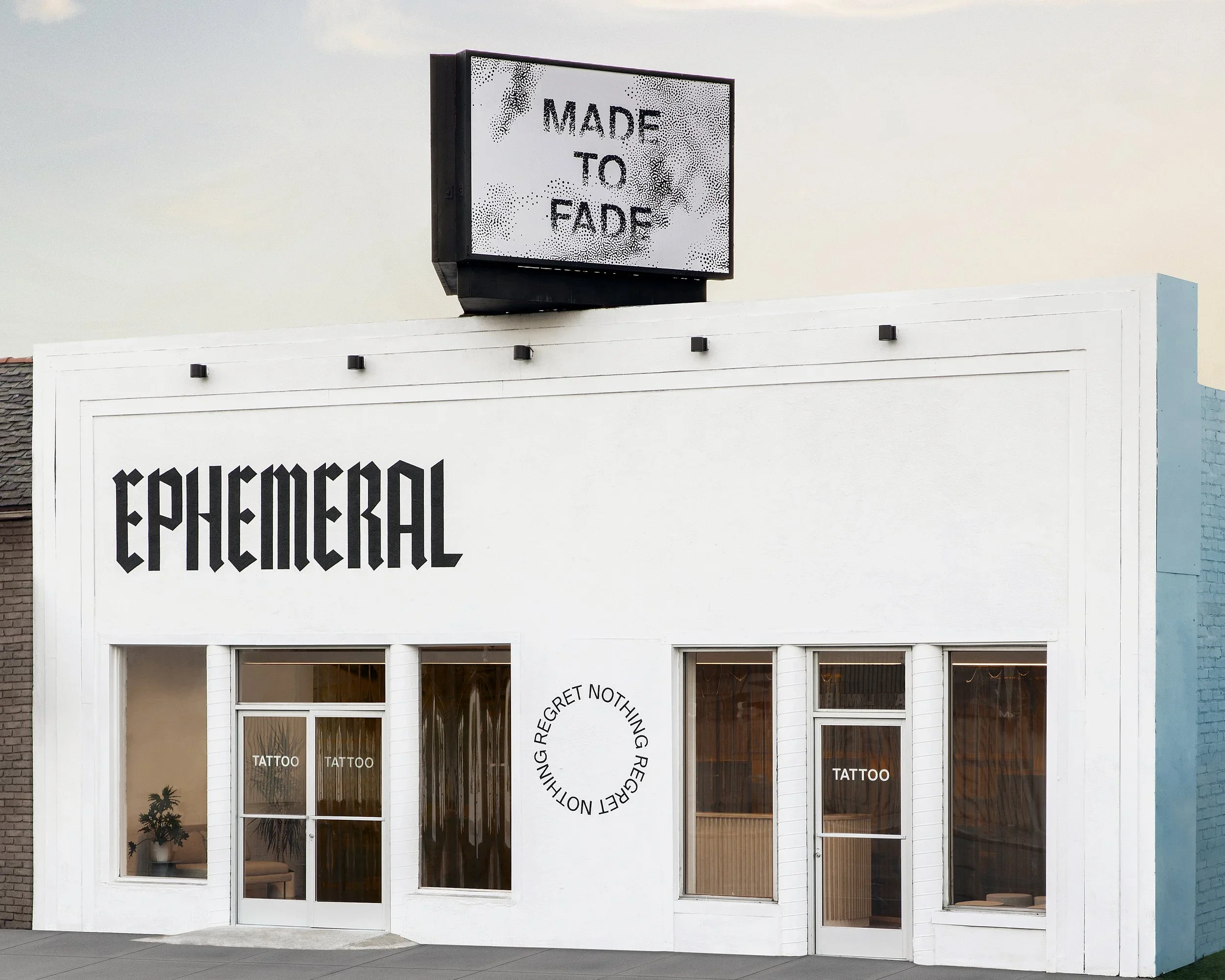 The storefront of a tattoo shop with large windows displaying decorations and furniture inside. A white wall features black text reading 'EPHEMERAL' and a circular design with words, and there is a black and white sign on the roof that says 'MADE TO FADE'.