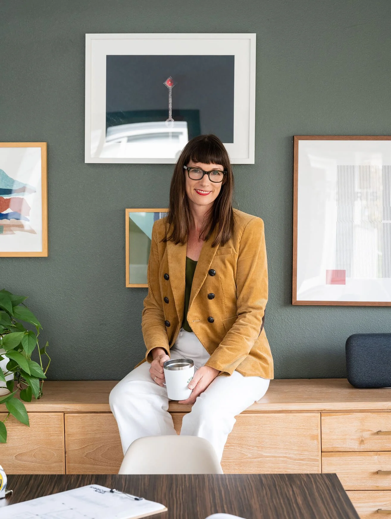 A woman with glasses and wearing a mustard-colored blazer, sitting on a wooden bench in a room with green walls and framed artwork, holding a white mug with both hands and smiling at the camera.