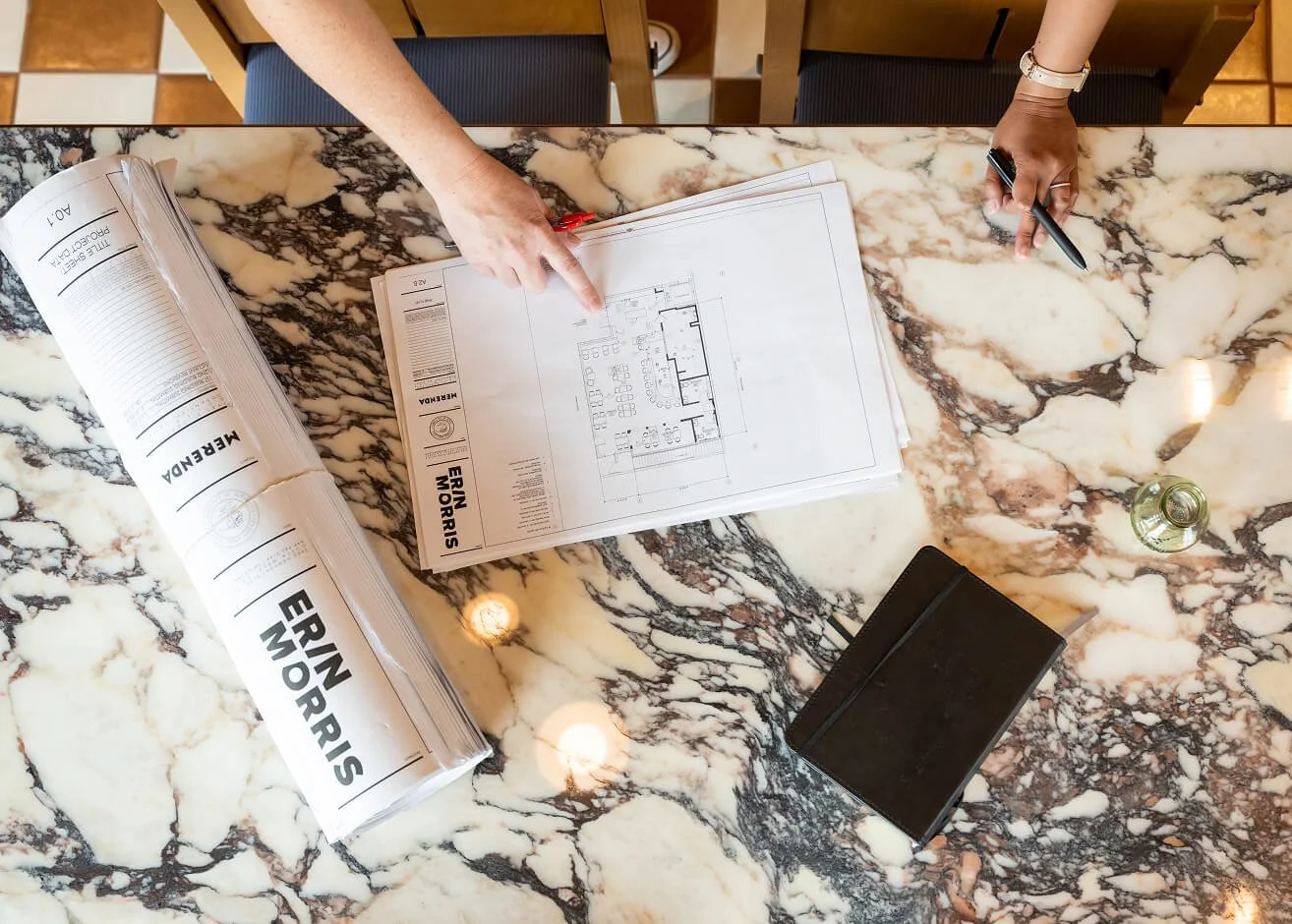 Overhead view of a marble table with architectural plans from 'Erin Morris', a rolled-up set labeled 'Merenda', a black notebook, a glass bottle, and two people pointing at the plans with pens.