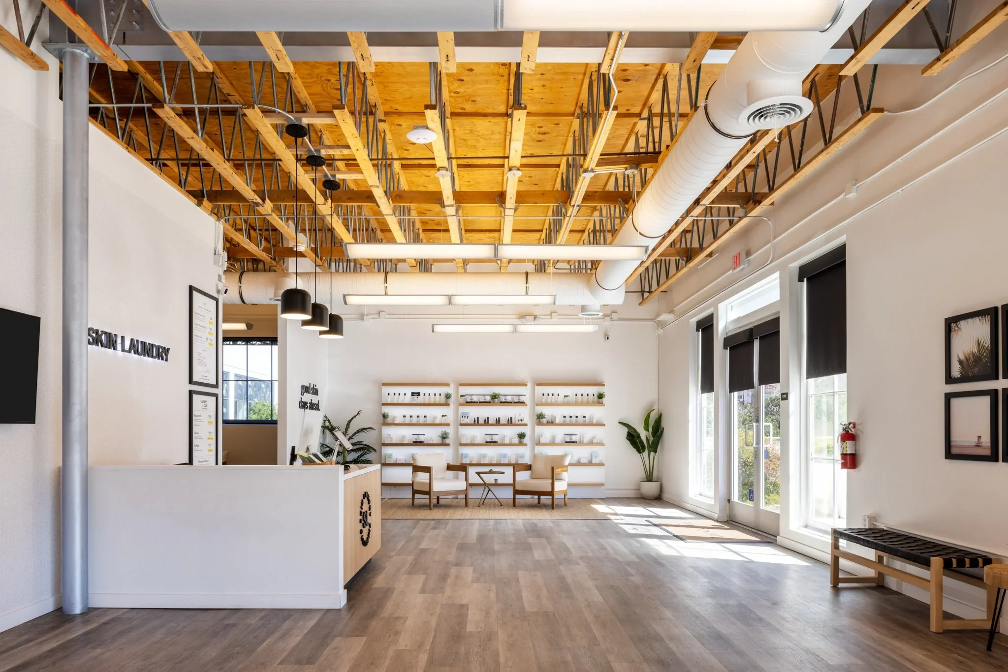 Interior of a modern, well-lit office or retail space with a wooden ceiling, exposed ductwork, and black pendant lights. Potted plants and framed artwork are on the white walls. There is a reception or information desk, seating area with chairs, and large windows letting in natural light.