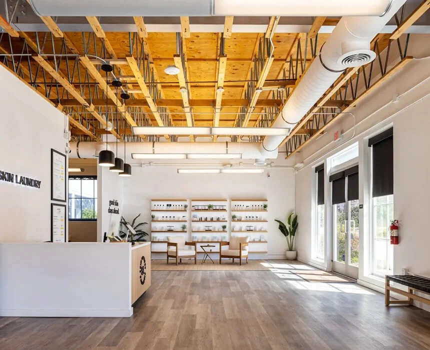 Modern, minimalist interior of a skin laundry clinic with wooden ceiling beams, white walls, large windows with black blinds, and a reception desk area with framed certificates. There are chairs and shelves with small plants and products, and a fire extinguisher near the windows.