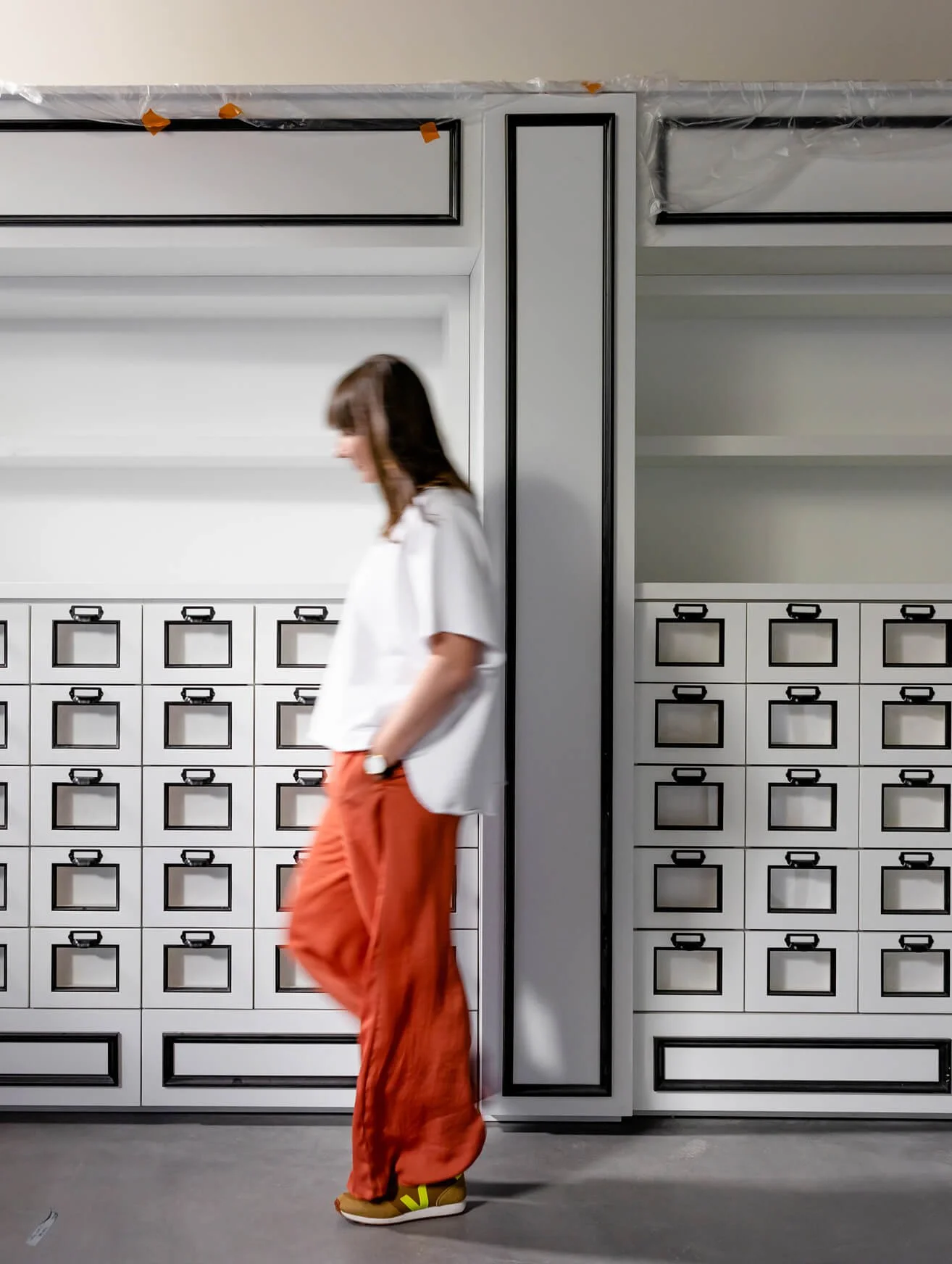 A woman wearing an orange pants and a white shirt walks past a large set of white storage lockers in an indoor setting.