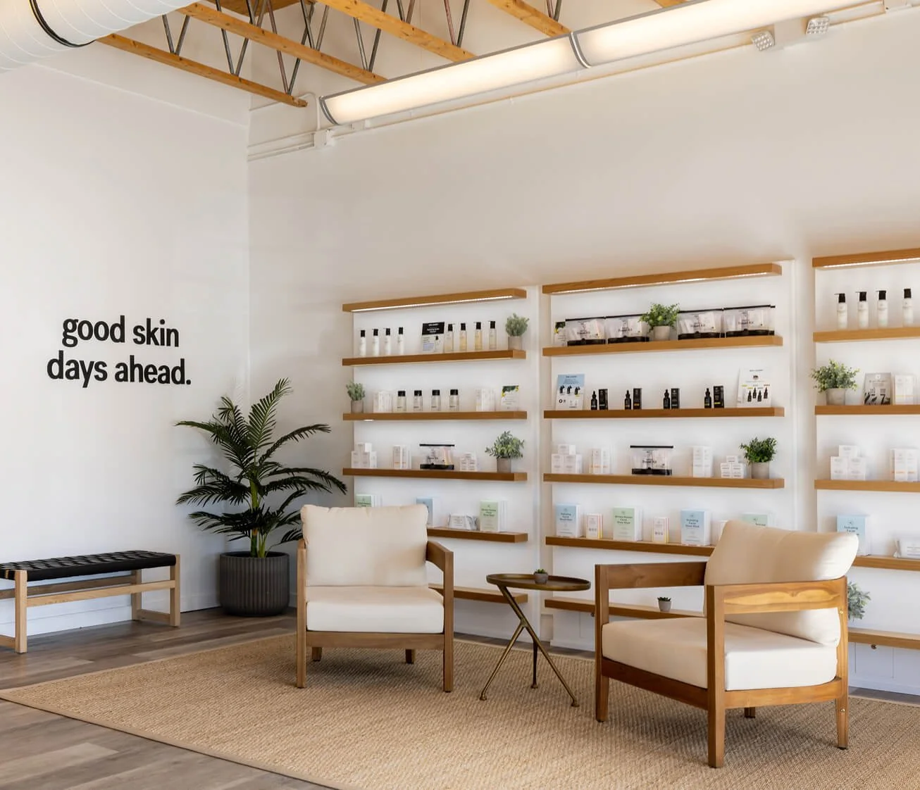 Minimalist waiting area with two beige armchairs, a small wooden side table, a potted plant, and a wall with shelves displaying skincare products, with the phrase 'good skin days ahead' on the wall.