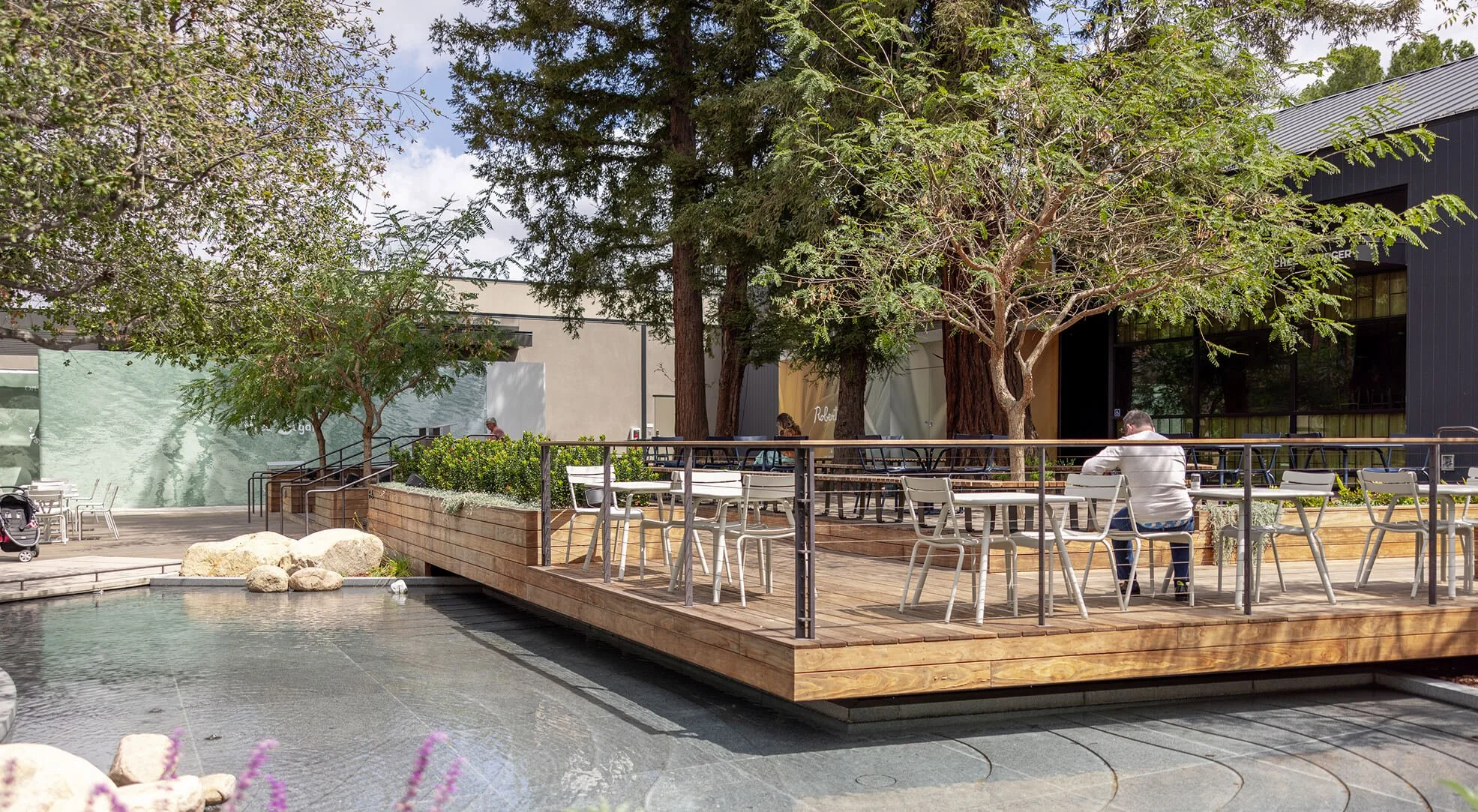 Outdoor seating area with tables and chairs on a wooden deck, surrounded by trees and plants, adjacent to a modern gray building.