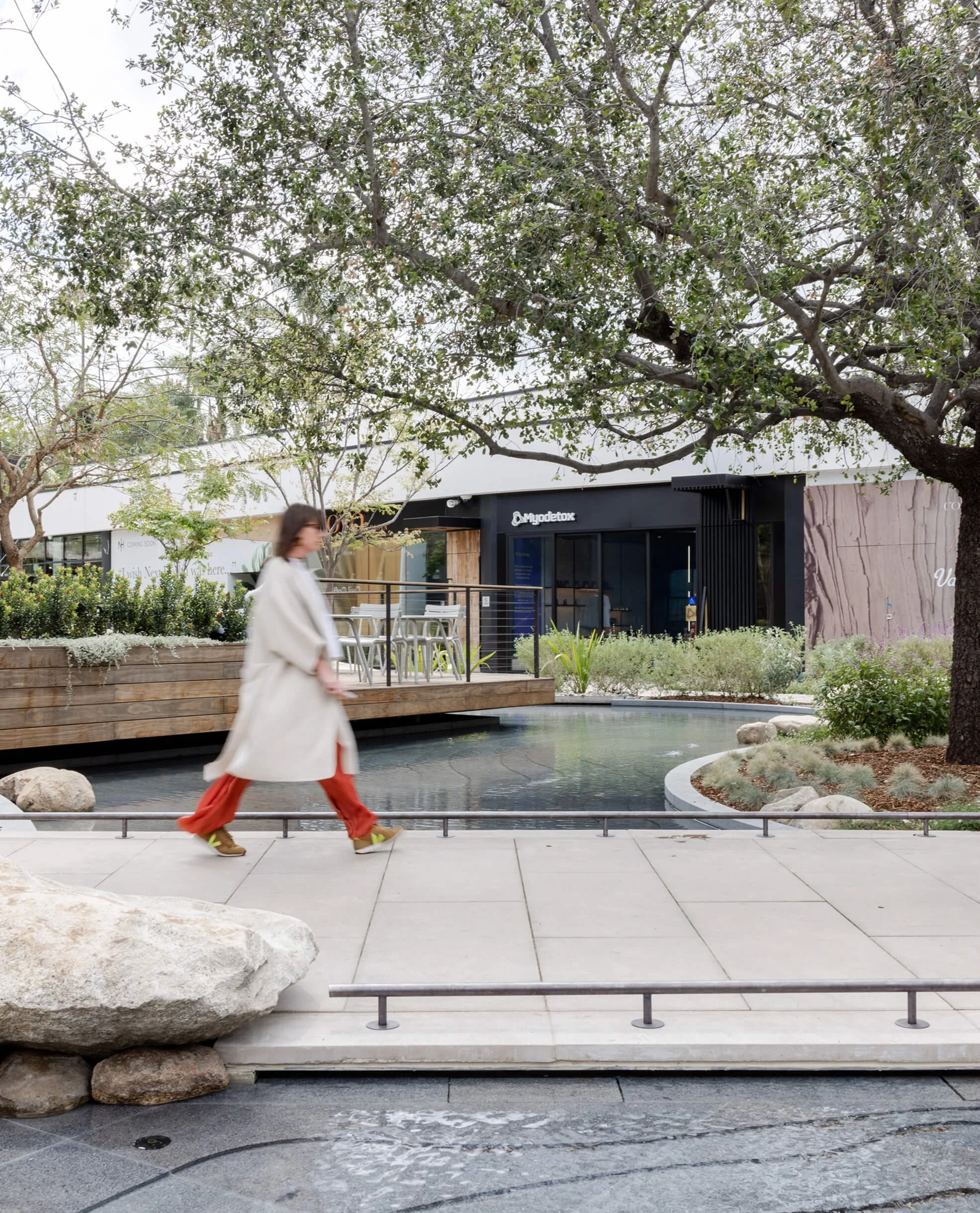 A person wearing a beige coat and orange pants walking past a modern building with the sign "Myodetox" in an outdoor urban setting with trees and landscaping.