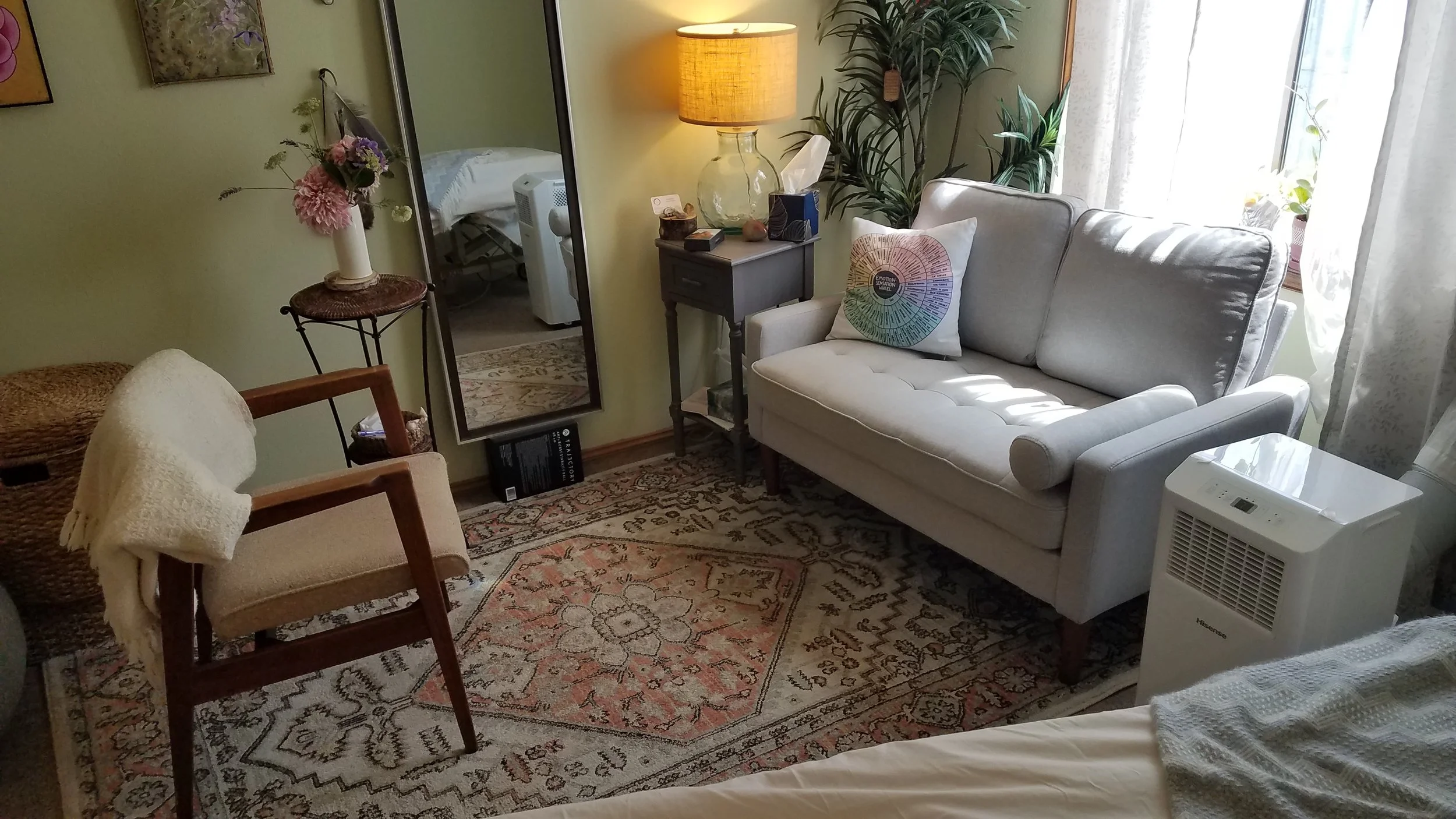 Joy Walker's office in Seattle with a white couch, a beige armchair with a blanket, a small wooden chair, a floral rug, a lamp on a side table, a large mirror, houseplants, and a window with sheer curtains.