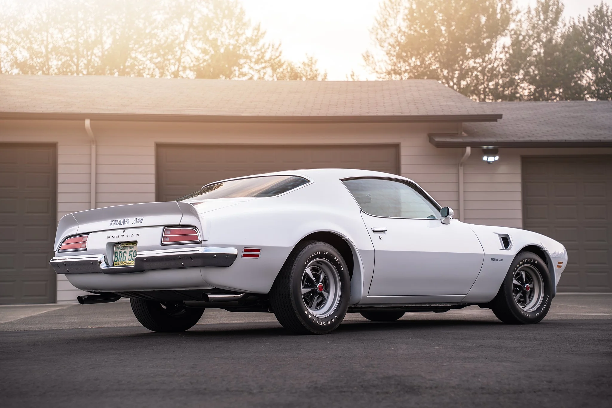 A classic white Pontiac Trans Am muscle car parked in front of a garage with closed doors, with sunlight filtering through trees in the background.