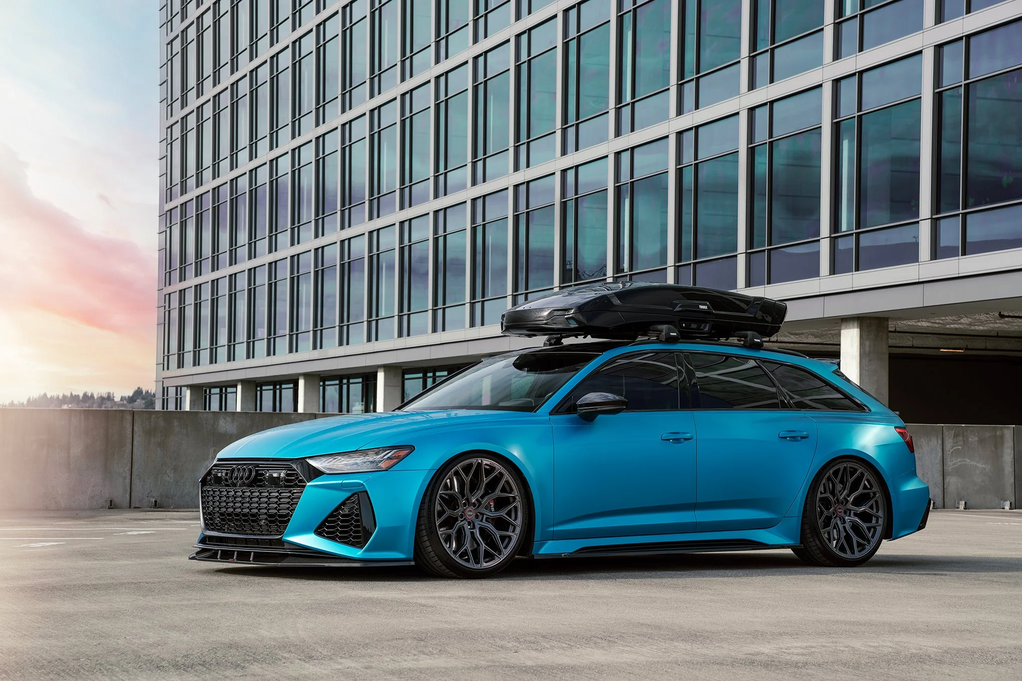 A blue Audi station wagon with a black roof box parked on a rooftop parking lot against a modern glass building at sunset.