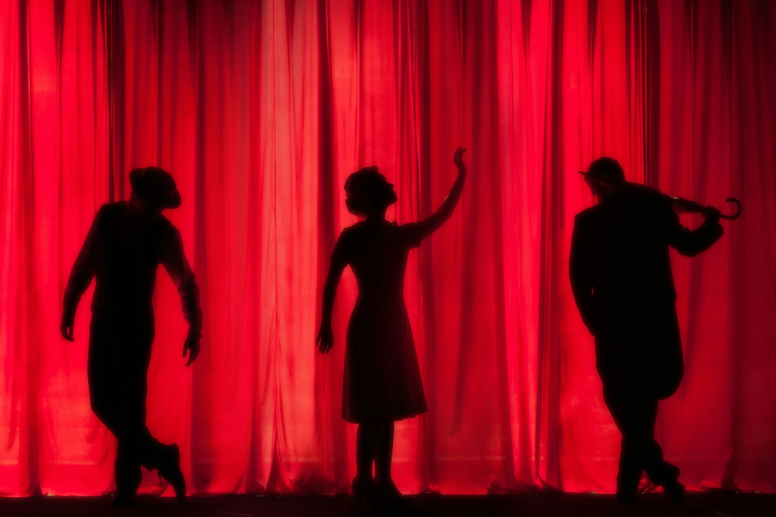 Silhouettes of three performers on stage in front of a red curtain, one holding a cane, one with hands raised, and the third leaning Musical Theater Dance Camp