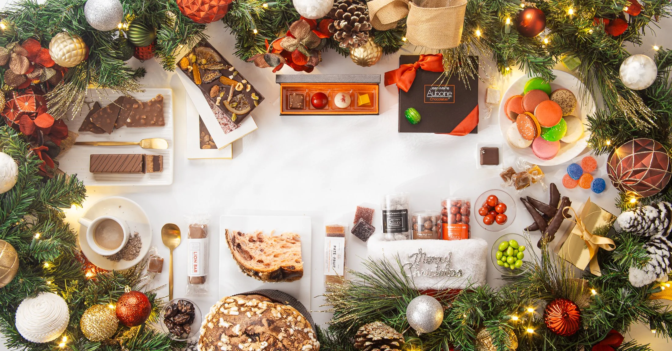 Luxury holiday flat-lay featuring premium chocolates, macarons, panettone, and gourmet treats framed by festive greenery