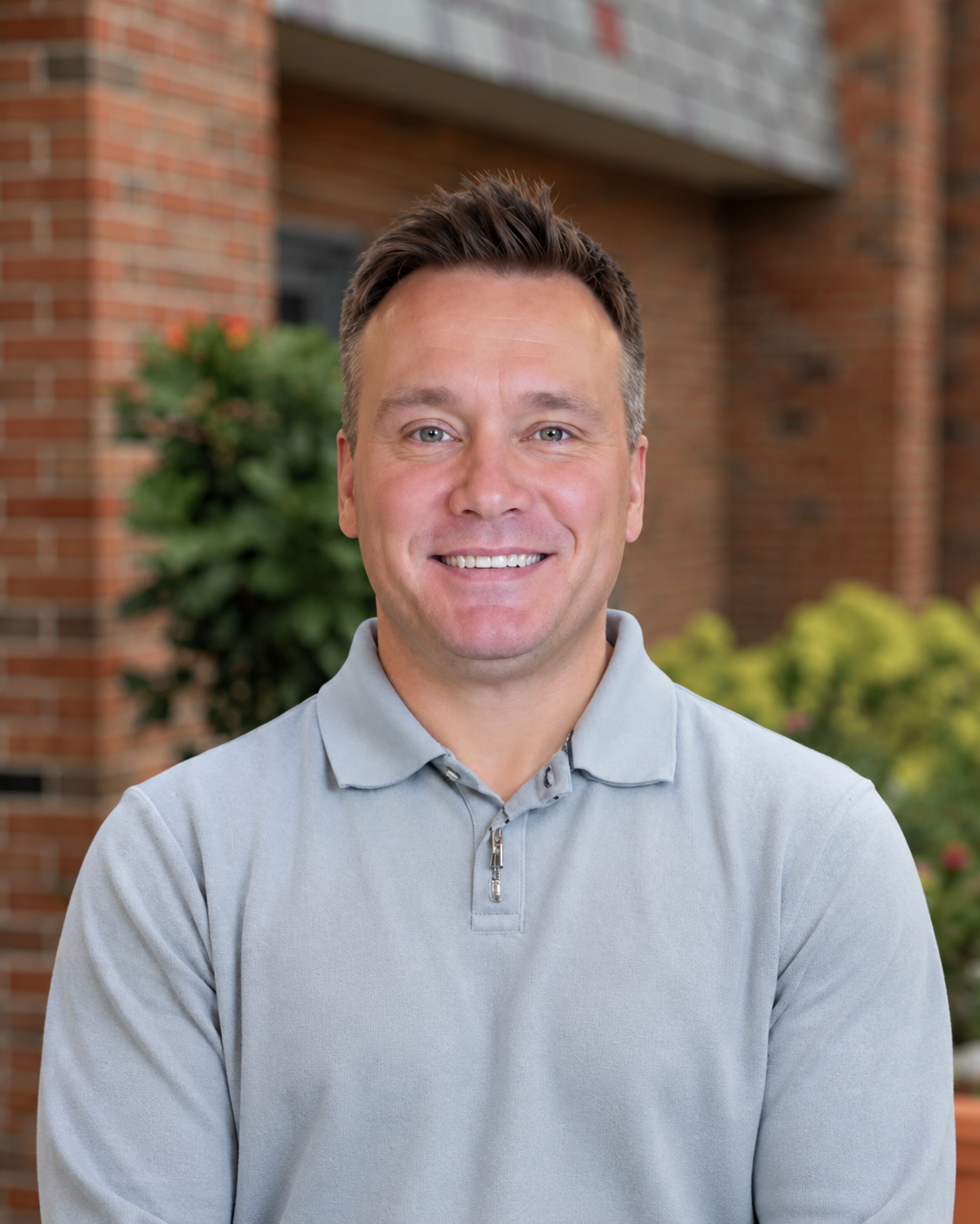 A man smiling outdoors in front of a brick building and green shrubbery, wearing a light gray collared shirt.