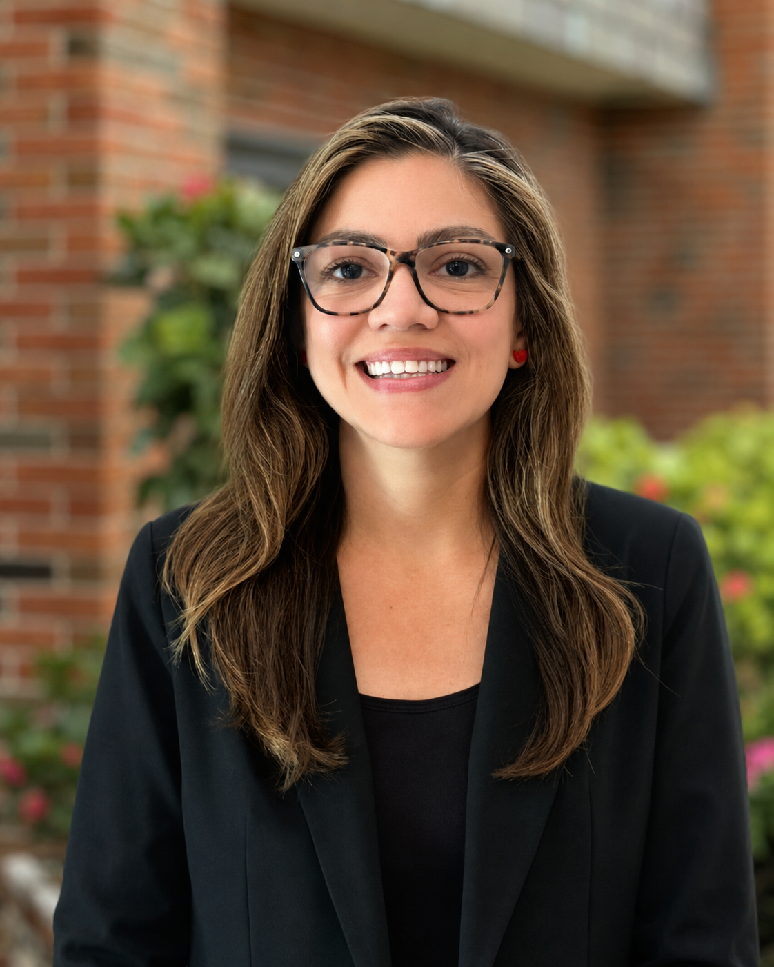 A young woman with long brown hair, glasses, and red earrings, smiling, outdoors in front of a brick building and greenery.
