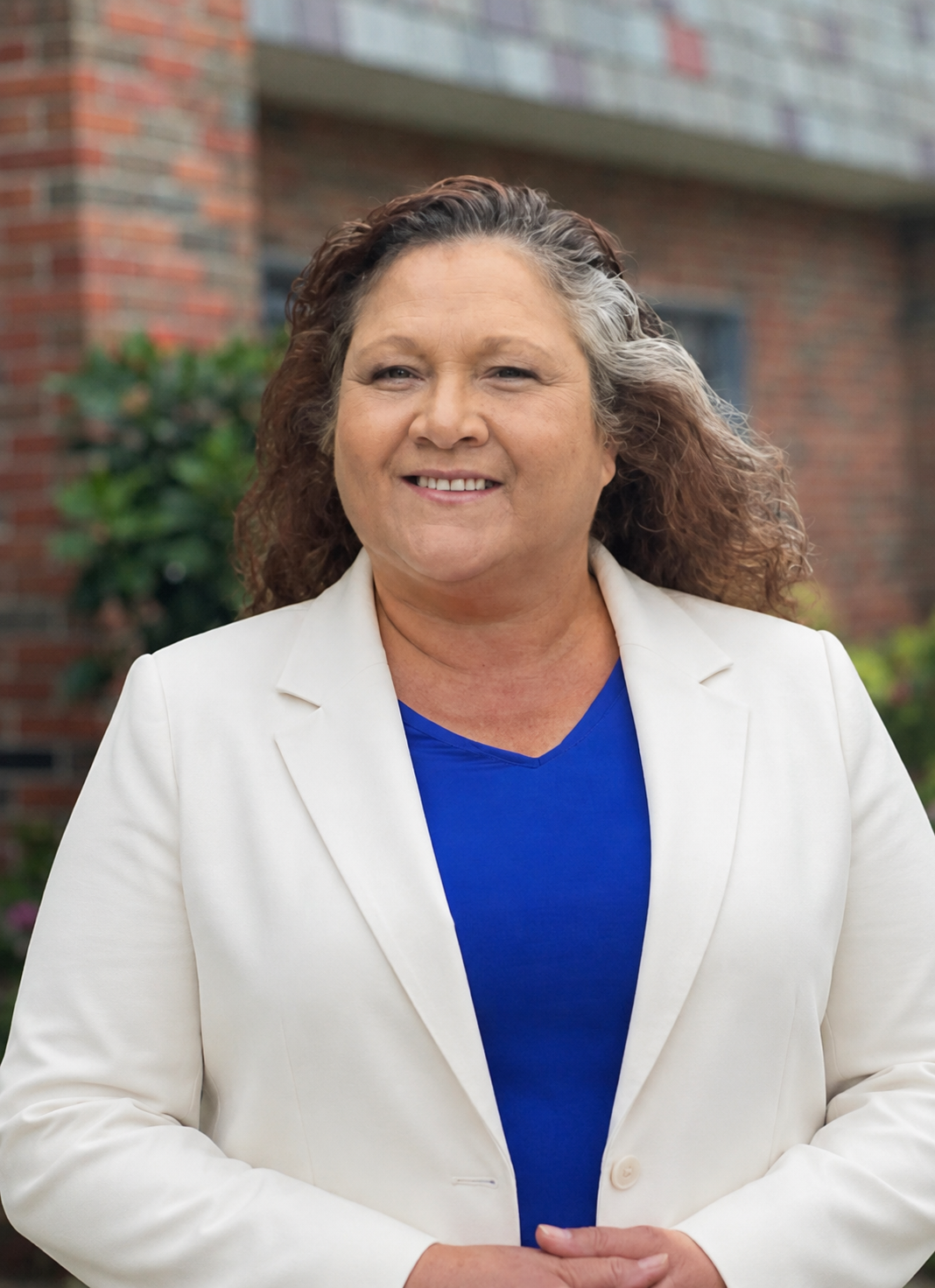 A woman with curly brown and gray hair smiling outdoors in front of a brick building and flowering bushes, wearing a white lace jacket over a blue top.