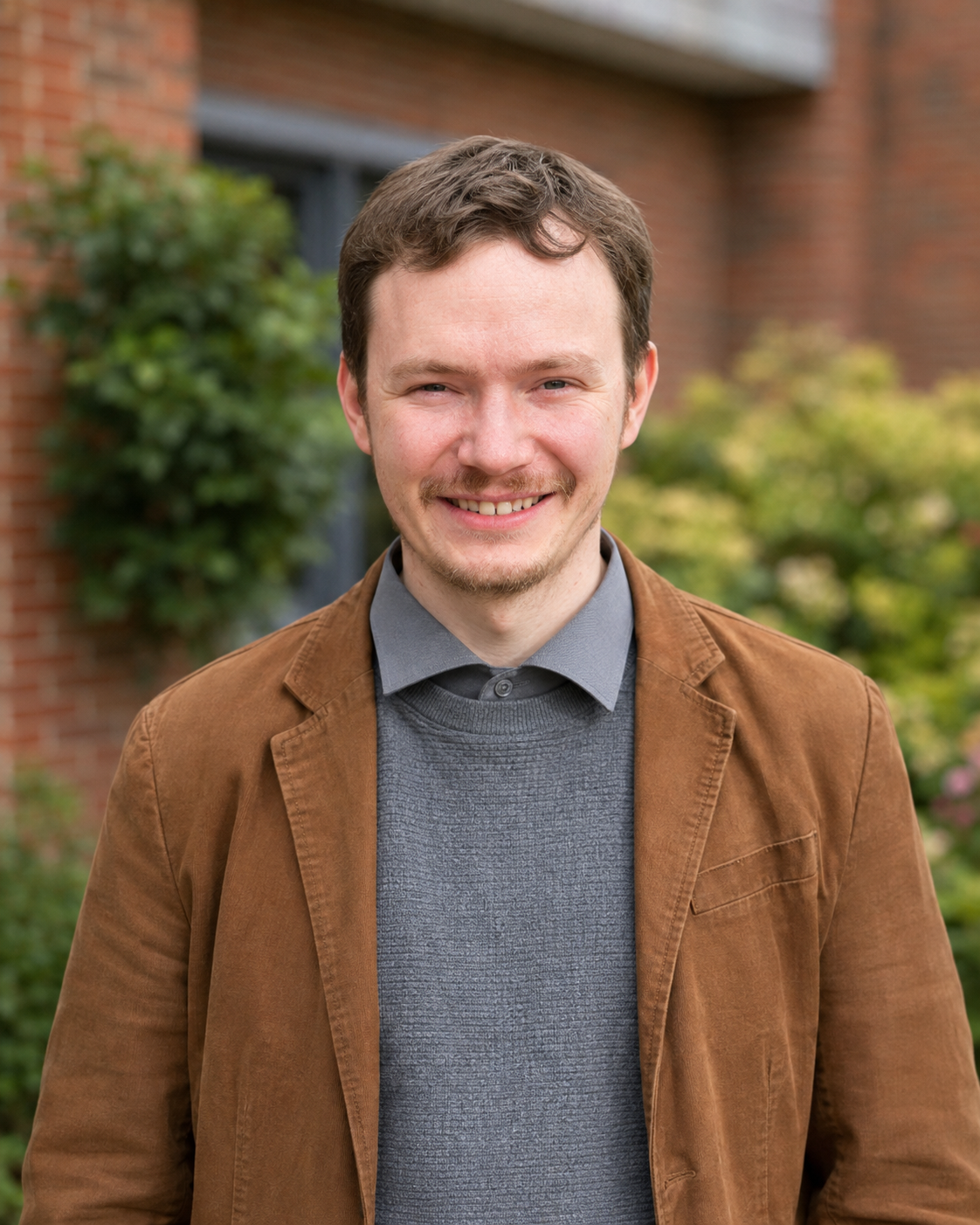 Portrait of a young man with brown hair, smiling, standing outdoors in front of a brick building and green bushes.