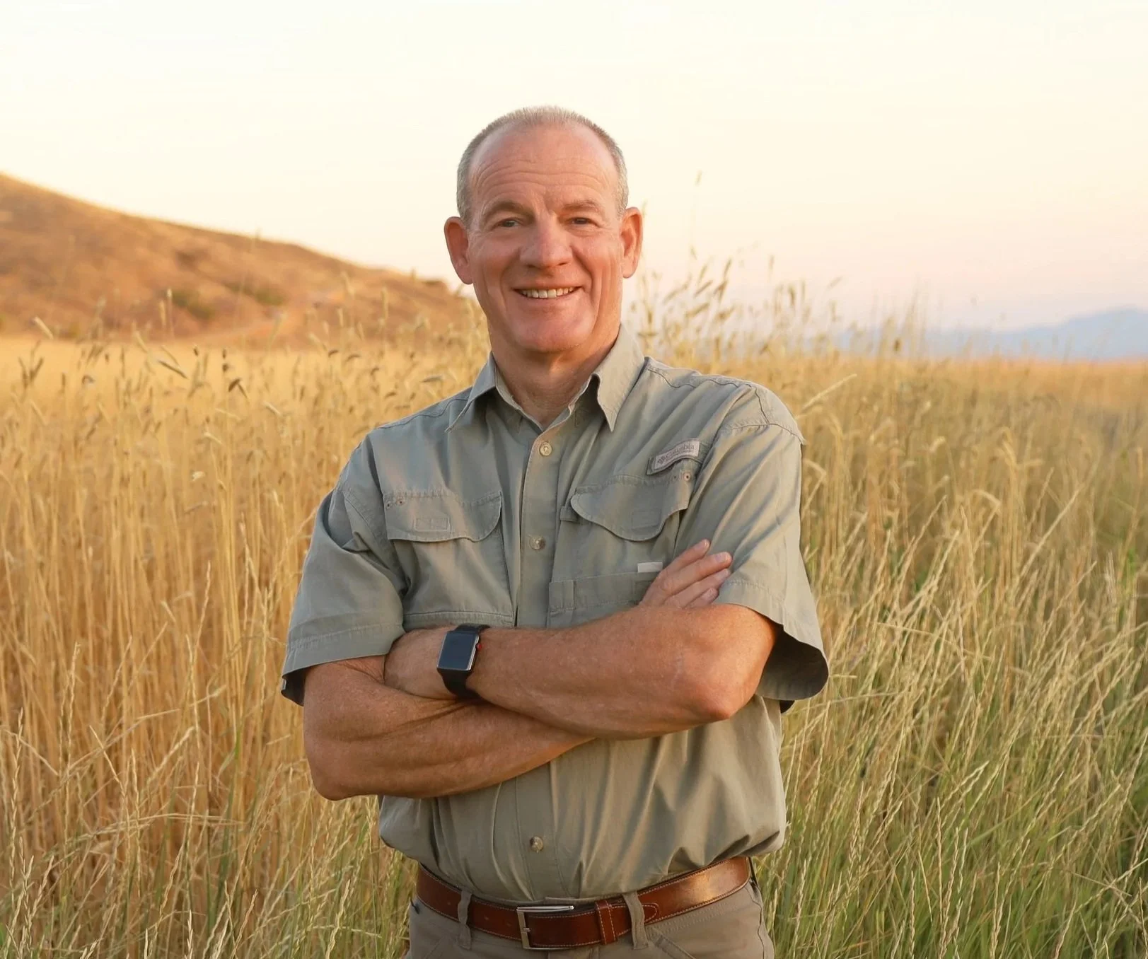 A smiling man outdoors in a grassy field, wearing a beige Columbia shirt and a smartwatch, leaning against a wooden fence during autumn.