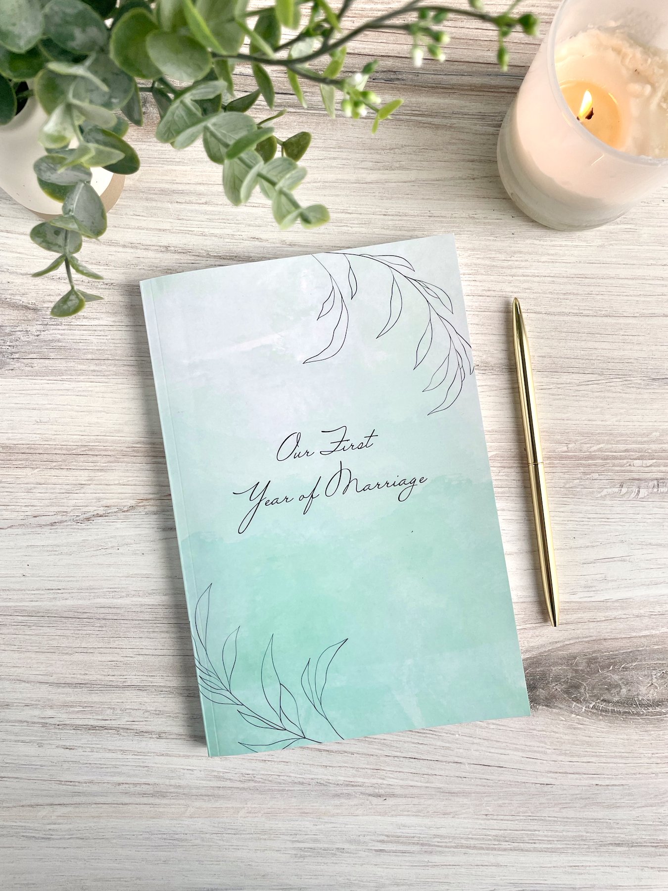 Our First Year of Marriage cover shown. Glossy softcover with light blue watercolor background and line-art leaves in the corners. A gold pen and white candle are visible.
