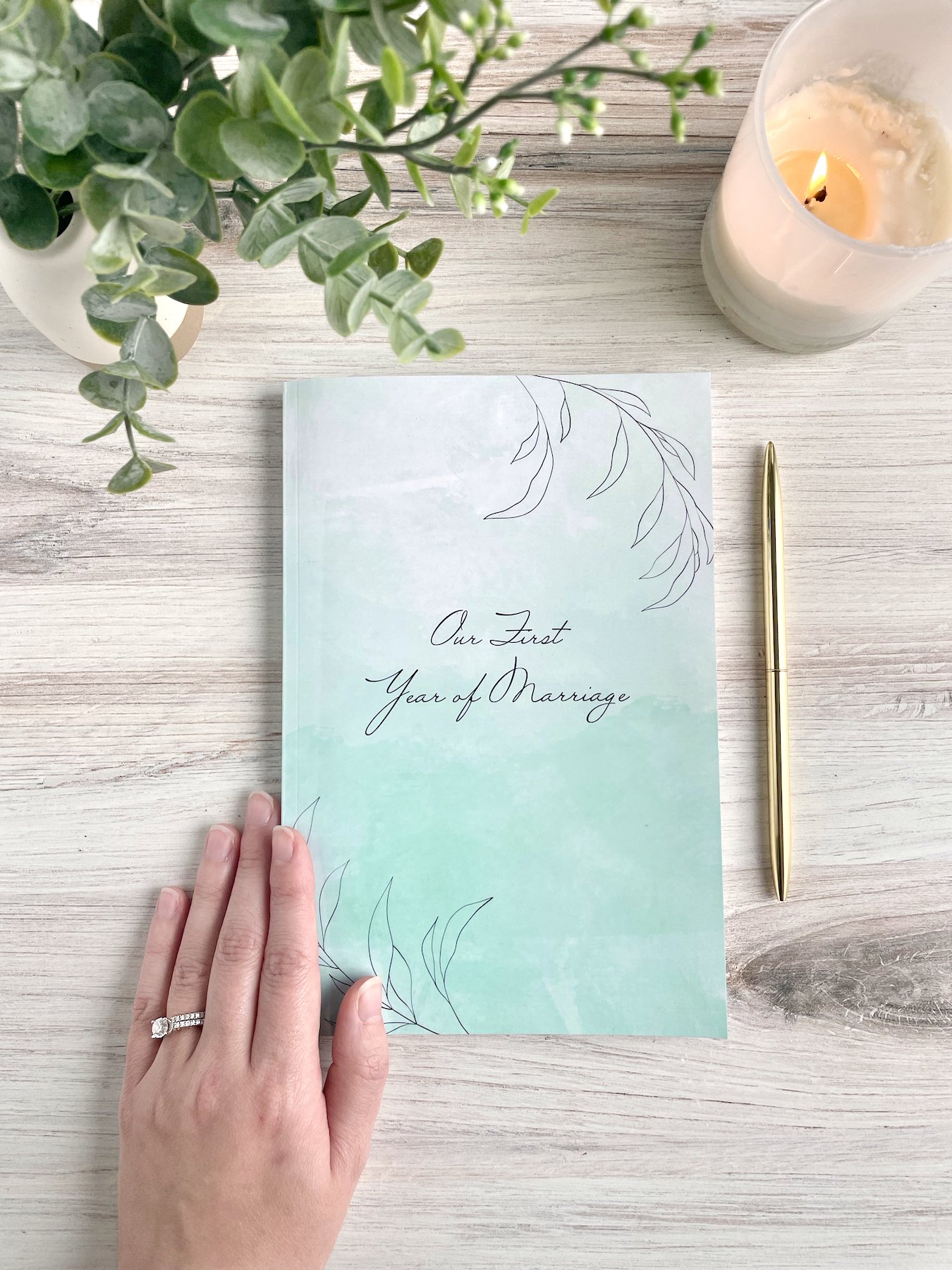Our First Year of Marriage cover shown. Glossy softcover with light blue watercolor background and line-art leaves in the corners. A gold pen and white candle are visible.