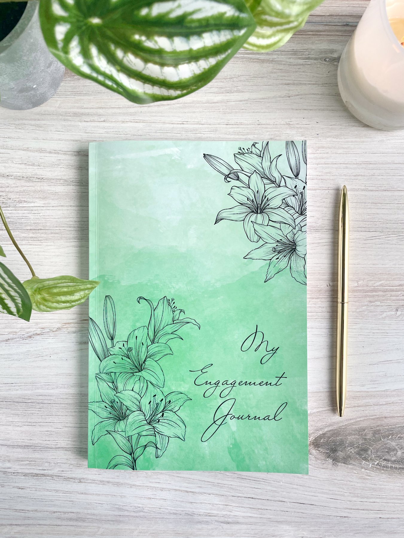 My Engagement Journal cover shown. Glossy soft-cover journal with green watercolor background and line-art lilies in the corners. A gold pen is visible.