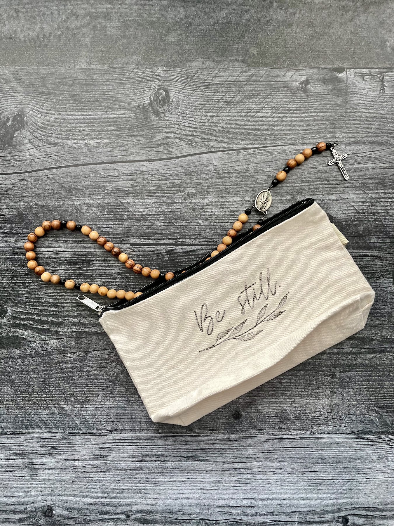 Canvas pouch with black zipper, stamped with cursive text "Be still." Pouch shows a wooden rosary.