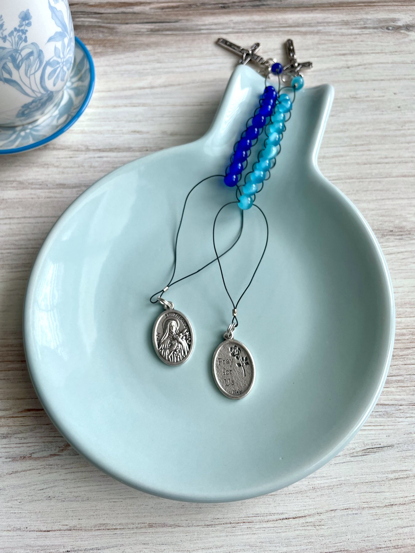 Two blue glass sacrifice beads are intertwined on a blue rest. The small silver St. Therese of Lisieux medals are shown, with St. Therese on one side and "Pray for us" on the other side.