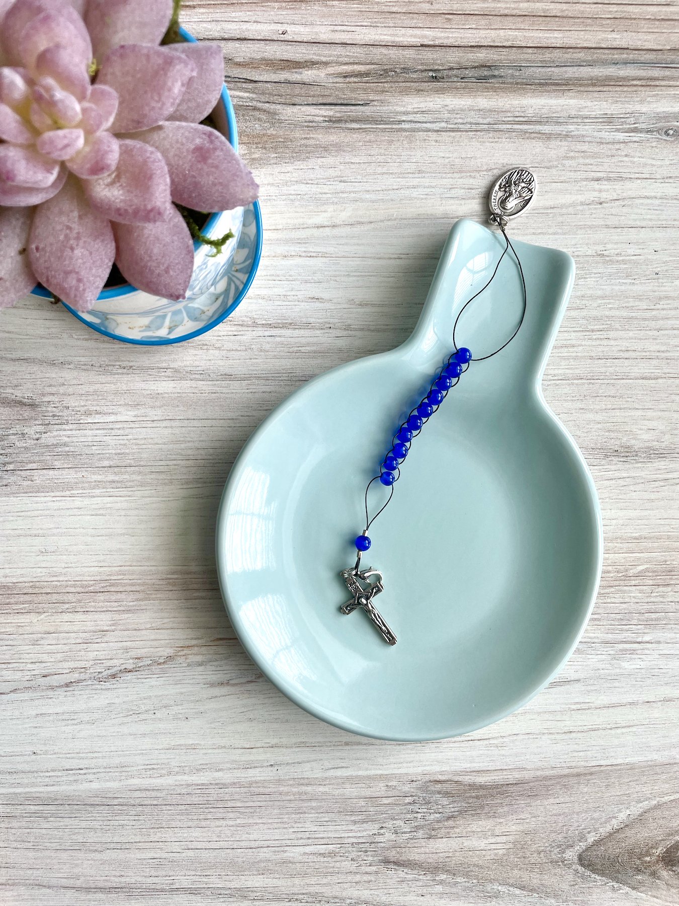 A dark blue glass sacrifice beads is resting in a blue dish. It includes a small silver crucifix and small silver St. Therese of Lisieux medal.