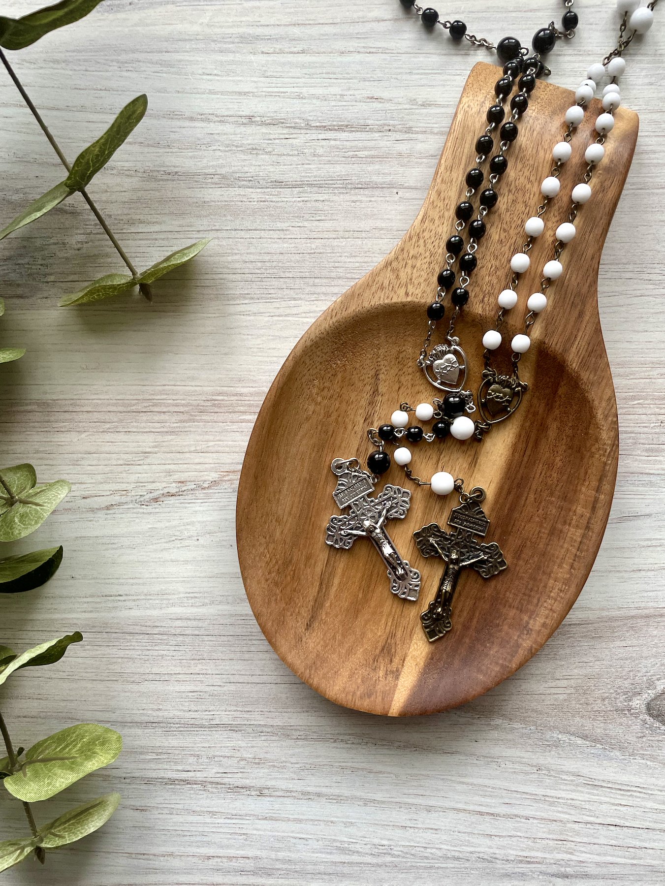 Black beaded rosary with silver chain, pardon crucifix, and Sacred Heart centerpiece on a wooden rest wrapped with matching white rosary with bronze chain, crucifix, and Sacred Heart centerpiece.