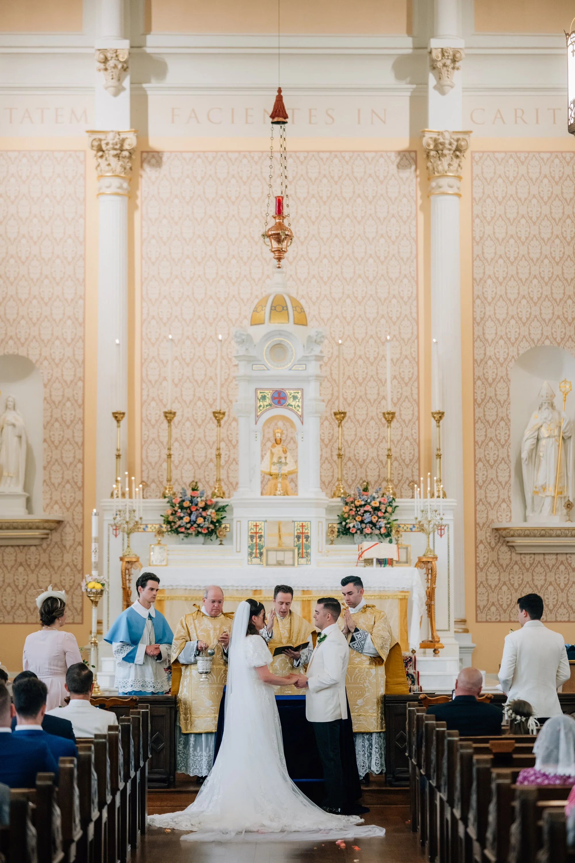 A bride and groom stand facing each other at the altar, saying their vows. A priest and several altar servers are behind them.