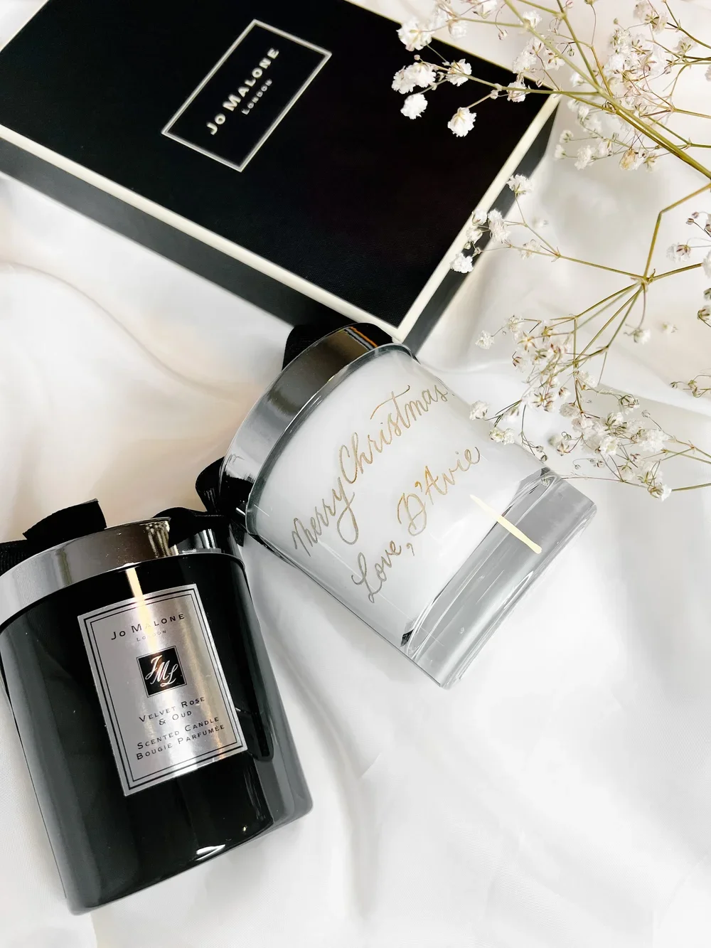 A gift set including two Jo Malone candles, a personalized glass candle holder with gold writing that says 'Merry Christmas Love, Daisie,' a black Jo Malone box, and white baby's breath flowers, all arranged on a white surface.