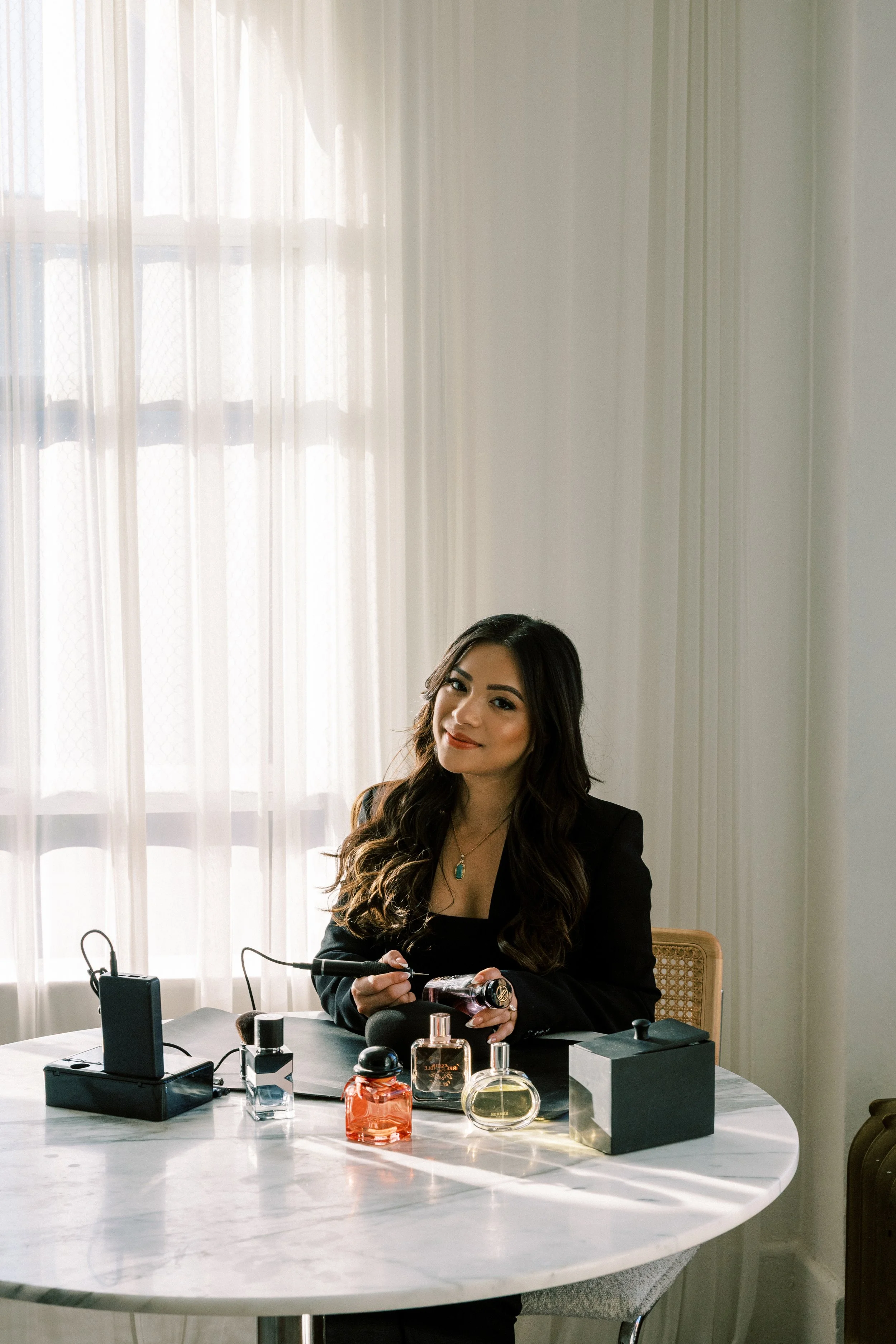 A woman with long dark hair and a black blazer sits at a white marble table with various perfume bottles, a black box, and a microphone, in a room with sheer curtains allowing natural light.
