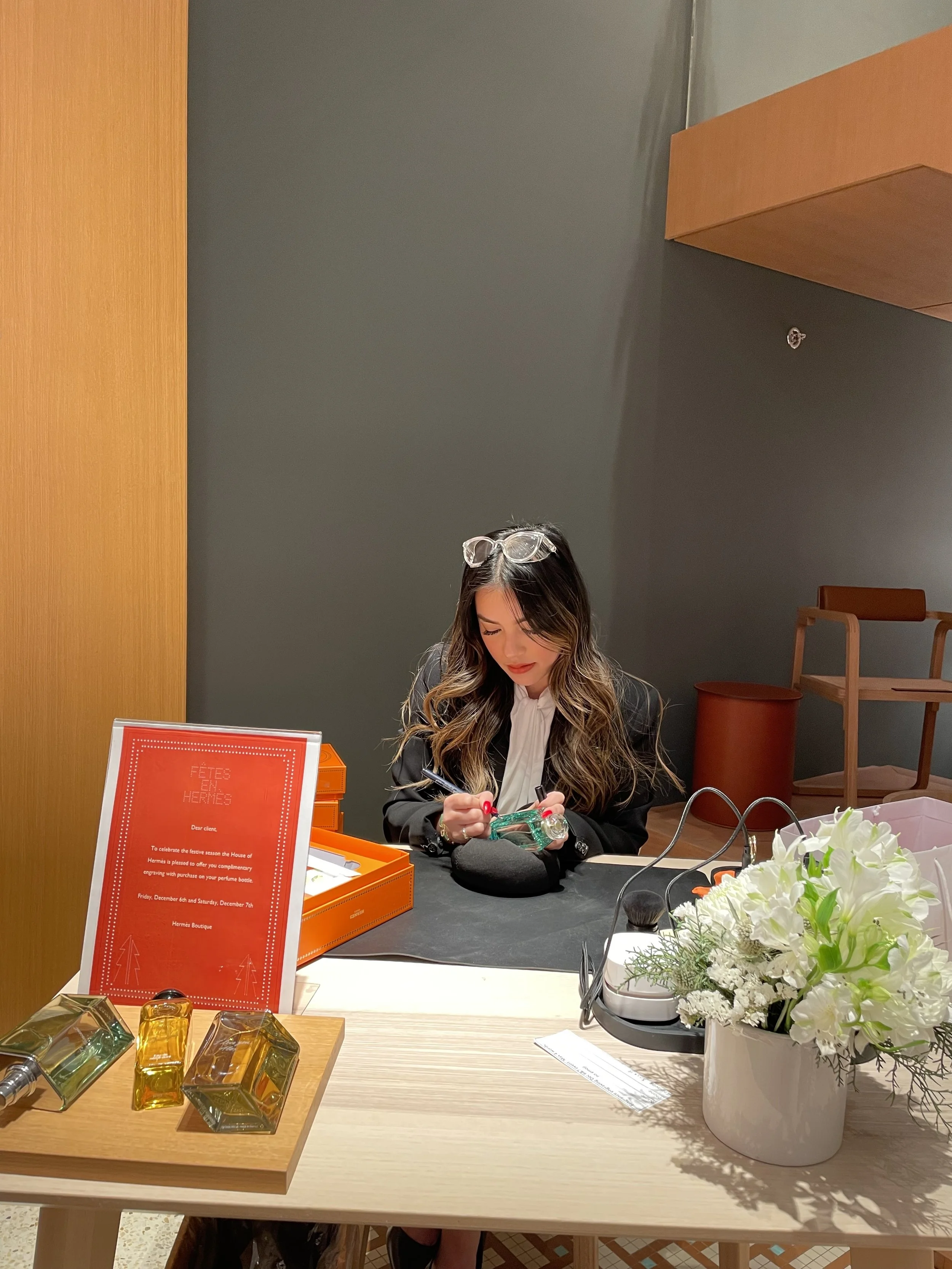 A woman with long wavy hair and glasses on her head is sitting at a table, holding a perfume bottle in her hands. The table has a sign, flowers, perfume bottles, and a black object. The background is a modern, minimalistic room with wooden and gray walls.