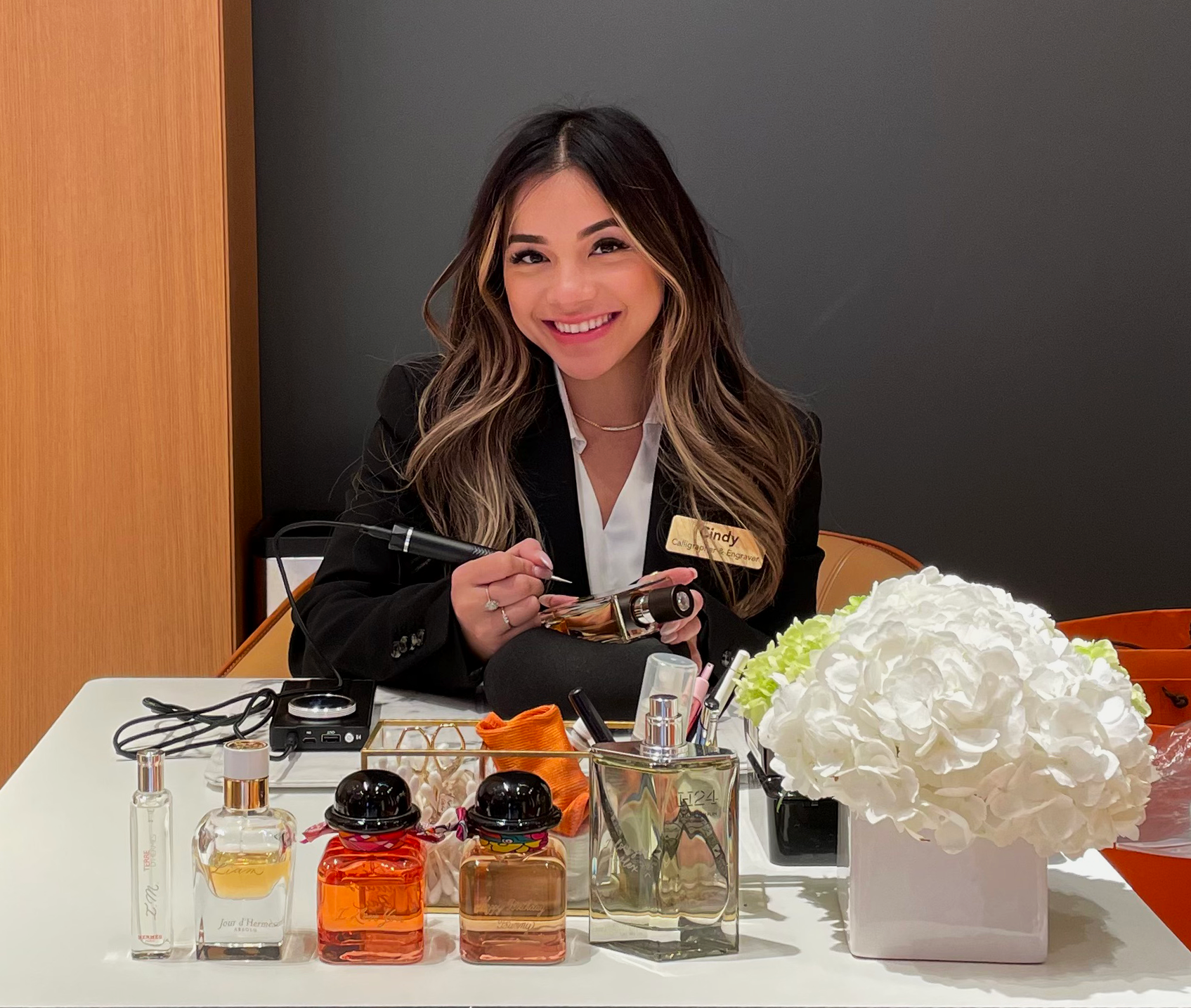 A woman with long wavy hair smiling at the camera, sitting at a table with perfume bottles and a bouquet of white flowers.