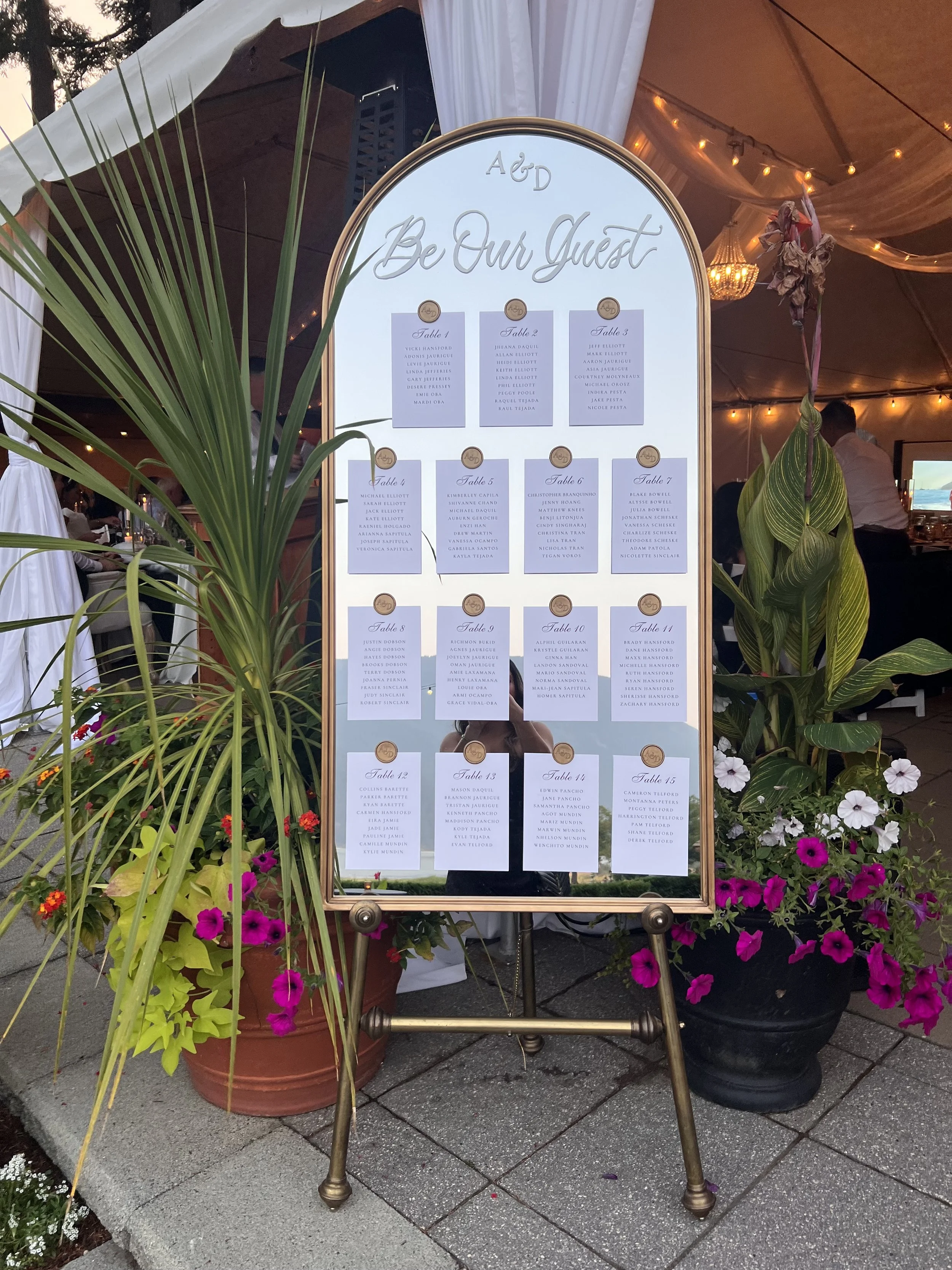 Wedding seating chart displayed on an easel board with list of guest names organized by tables, decorated with green plants and purple, pink, and white flowers, inside a tent with warm lighting.