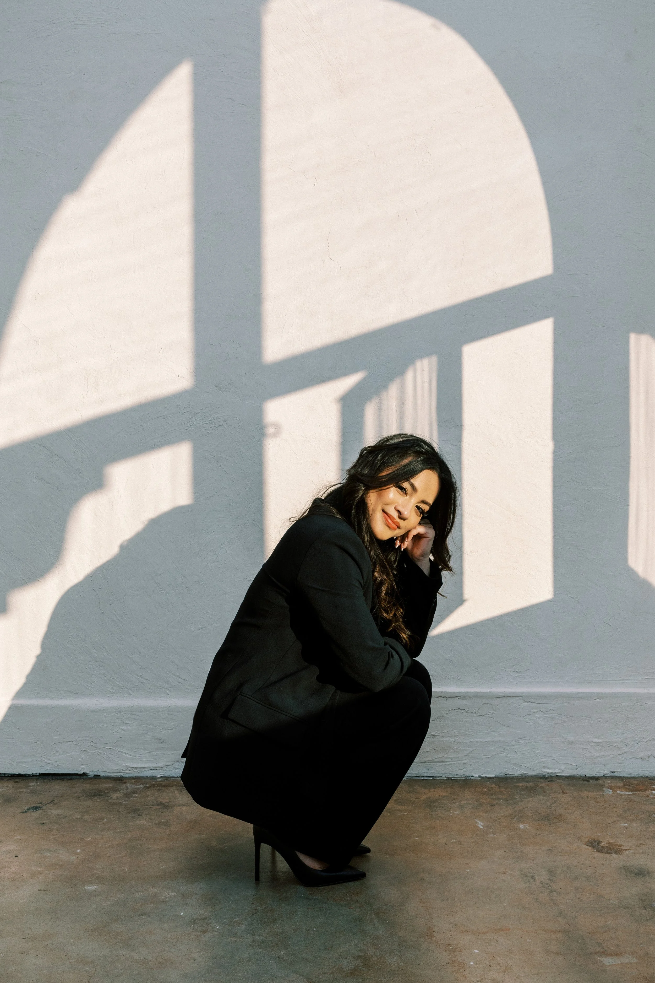A woman in a black suit squatting indoors, with sunlight casting window shadow patterns on the wall behind her.