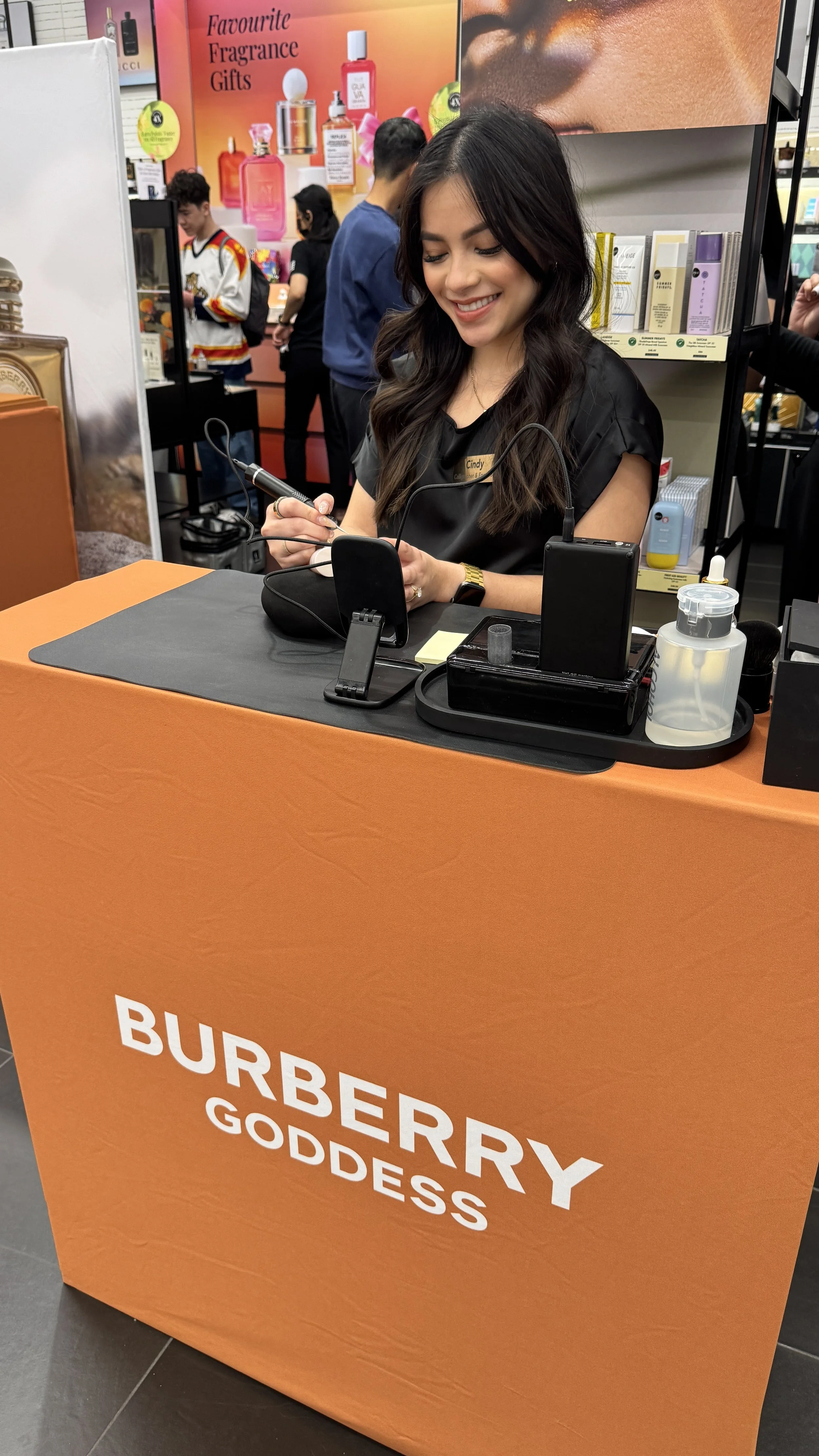 A woman smiling, sitting at a table with an orange cover that has the words 'BURBERRY GODDESS' on it, in a retail store. She is engaged in a demonstration with various tools and products on the table, and there are several people and product displays in the background.