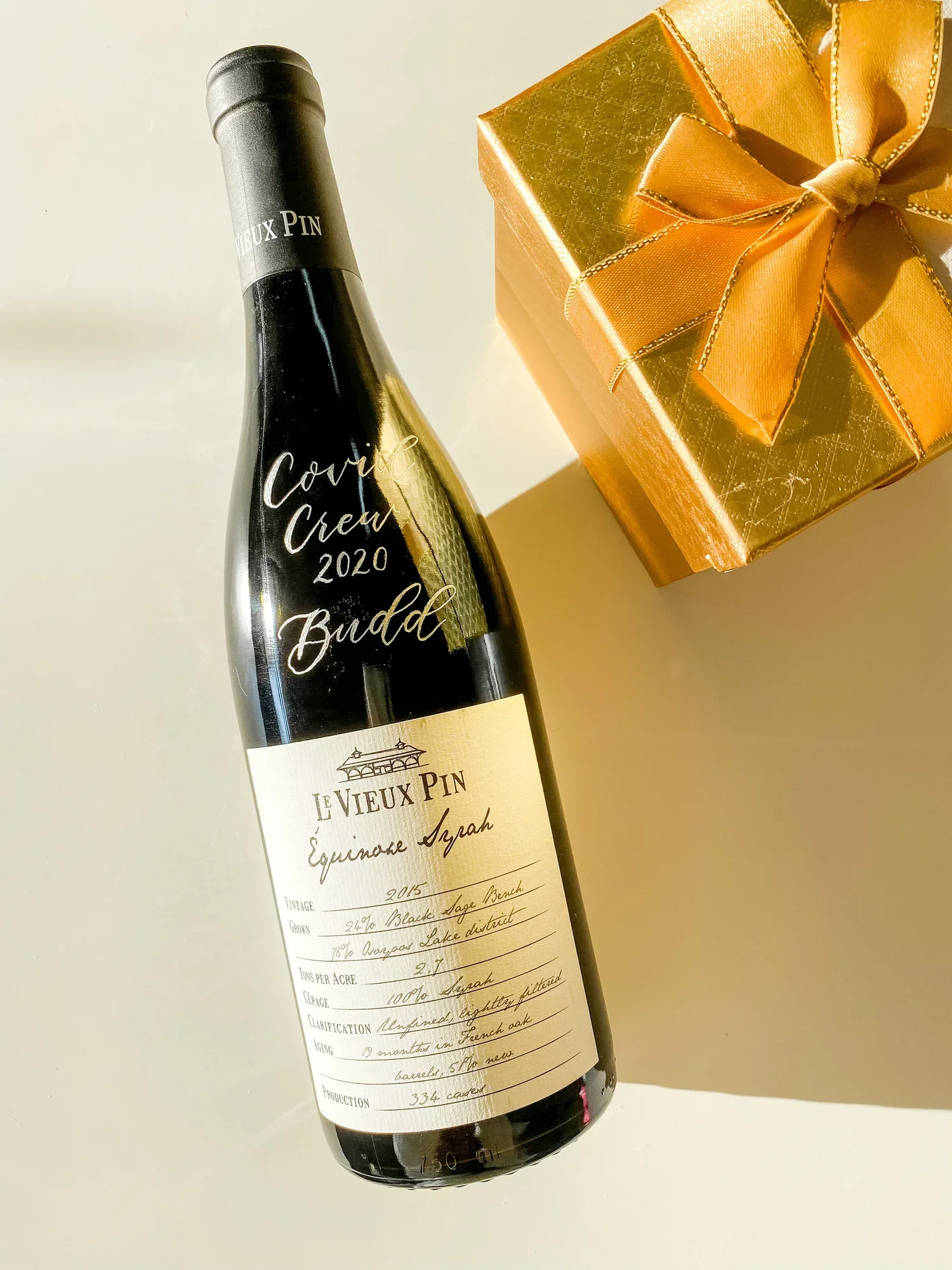 A personalized wine bottle labeled 'Covie Crew 2020' next to a gold gift box with a bow.