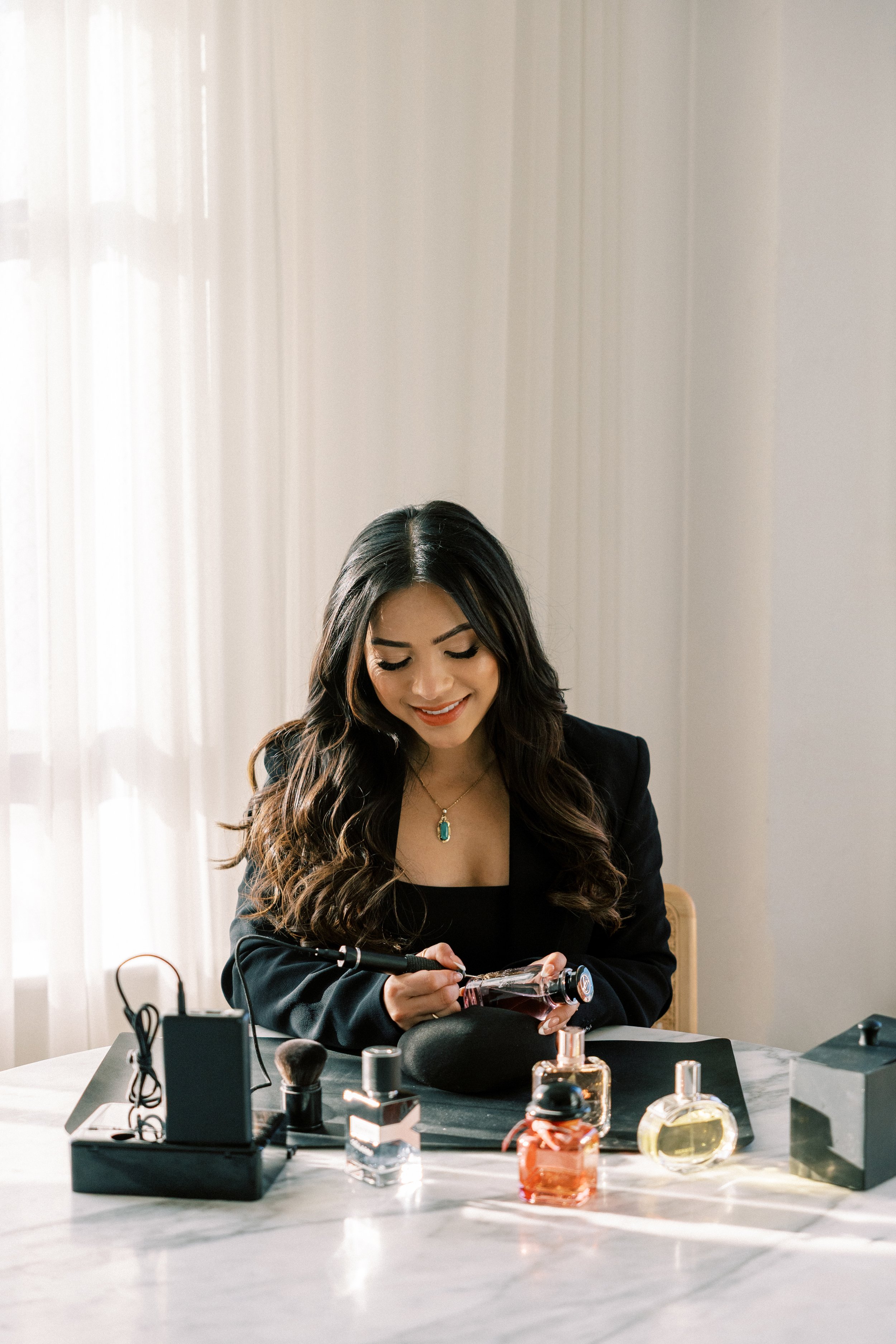 Woman in black blazer applying perfume to a perfume bottle at a table with various perfume bottles and a mirror in a bright room.