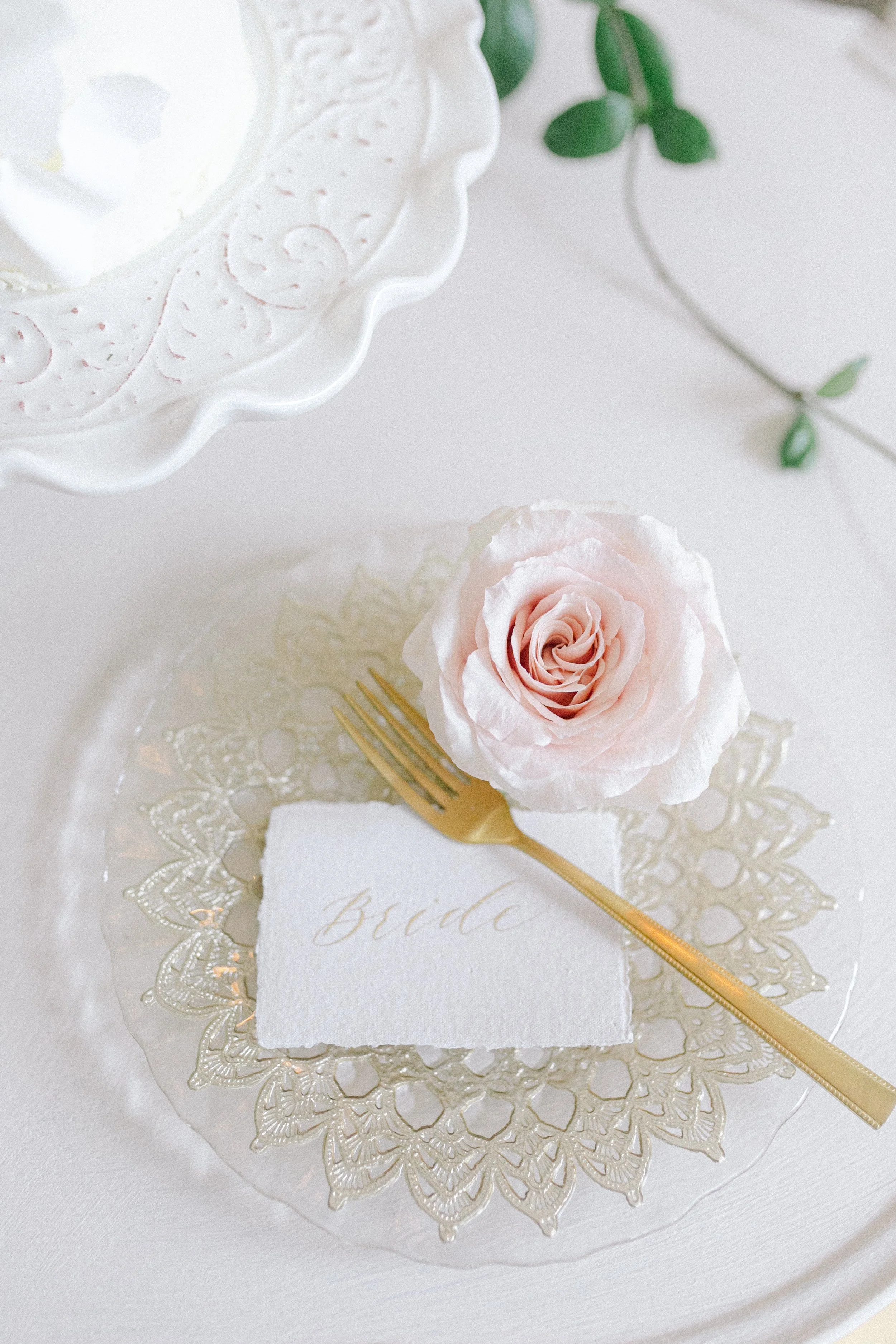 A pink rose on a lace-edged transparent plate with a white napkin labeled 'Bride,' a gold fork, and a white ornate cake stand in a soft, elegant setting.